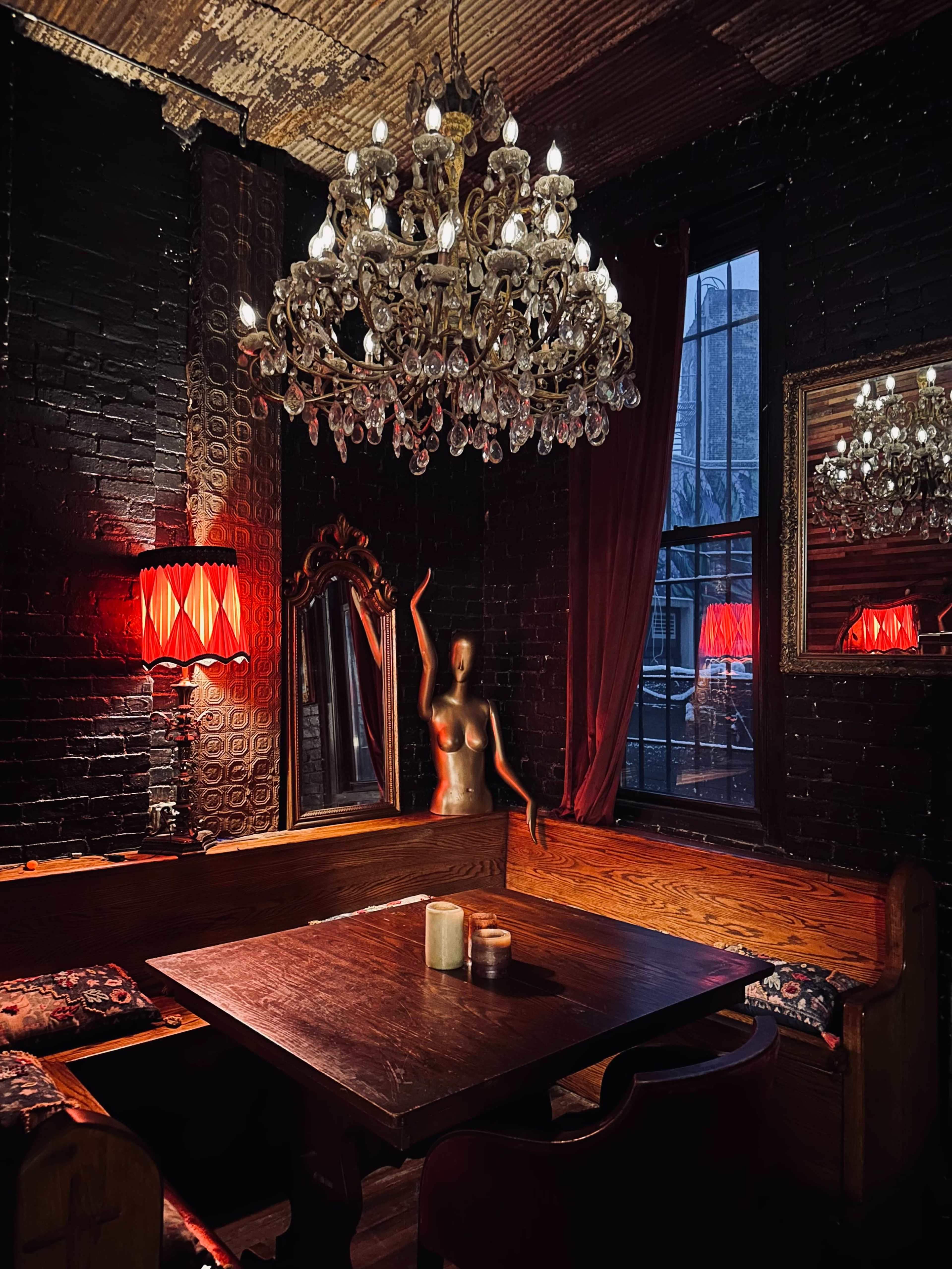 The image shows a dimly lit, cozy restaurant interior featuring a large chandelier, red lamps, a mirror, and a statue of a woman, with wooden seating around a dark table.
