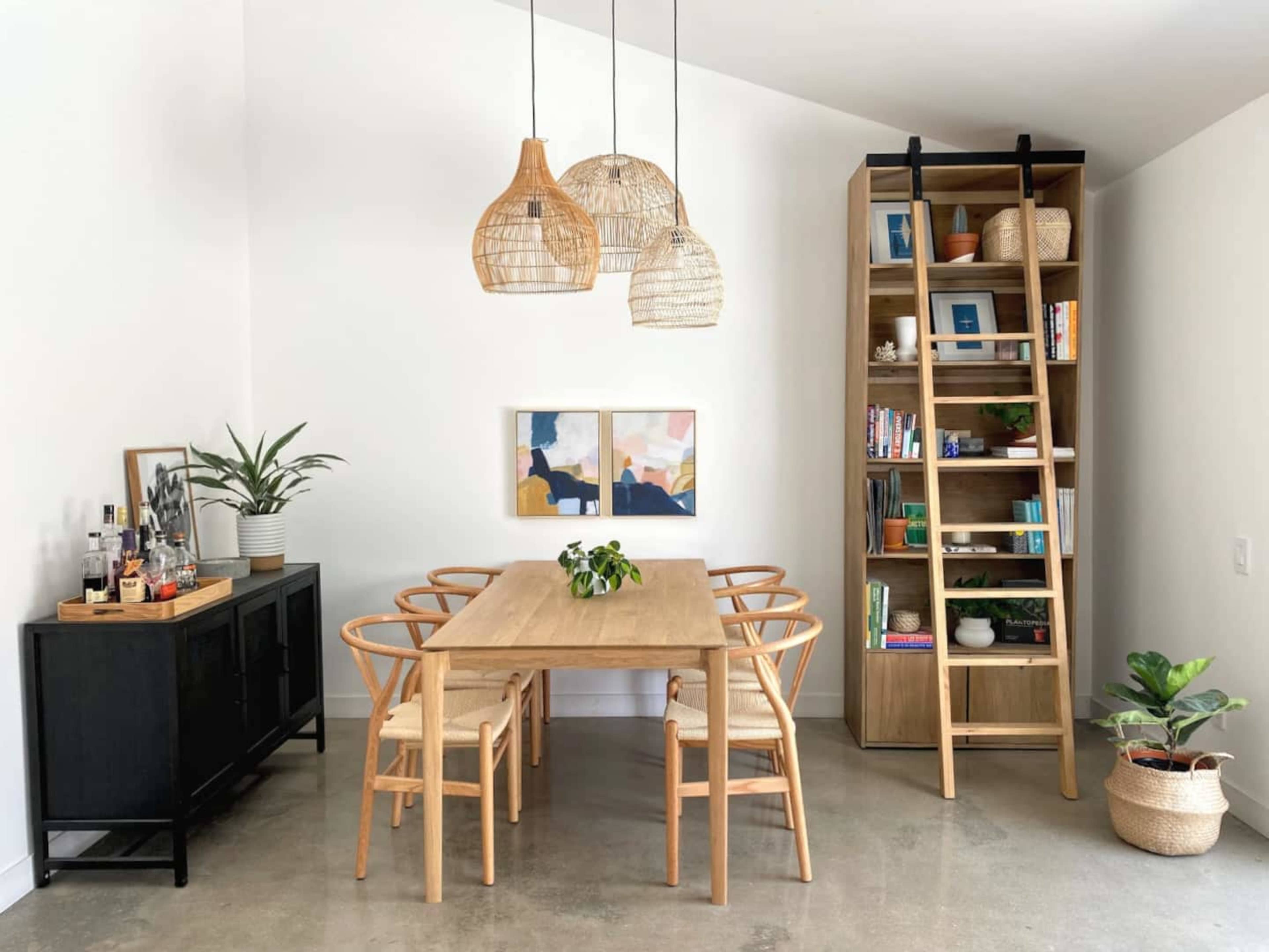 A modern dining area features a wooden table surrounded by six chairs, a tall bookshelf filled with books, and pendant lights hanging from the ceiling.