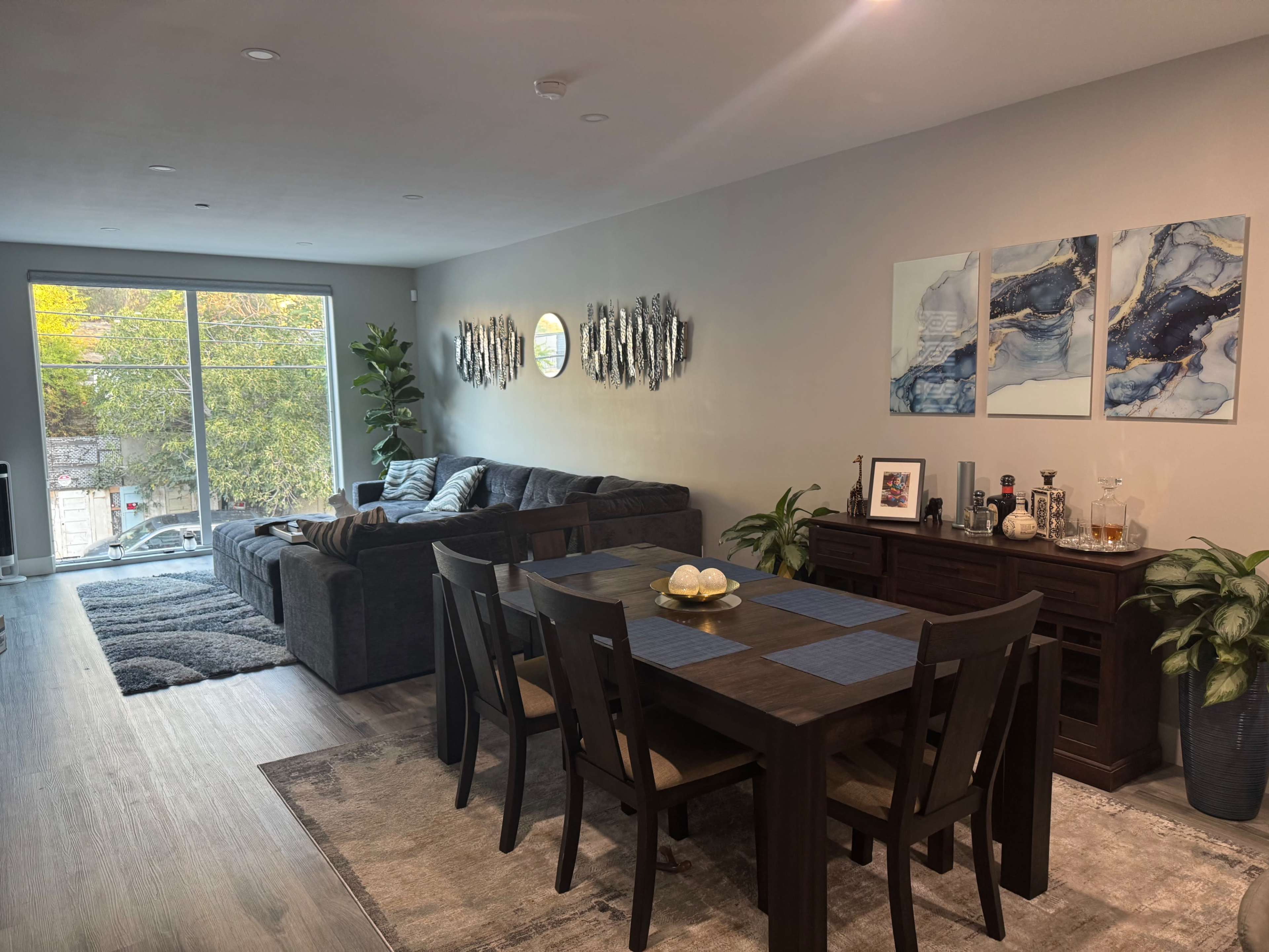 The image shows a modern living room and dining area featuring a gray sectional sofa, a wooden dining table with chairs, and decorative artwork on the walls.