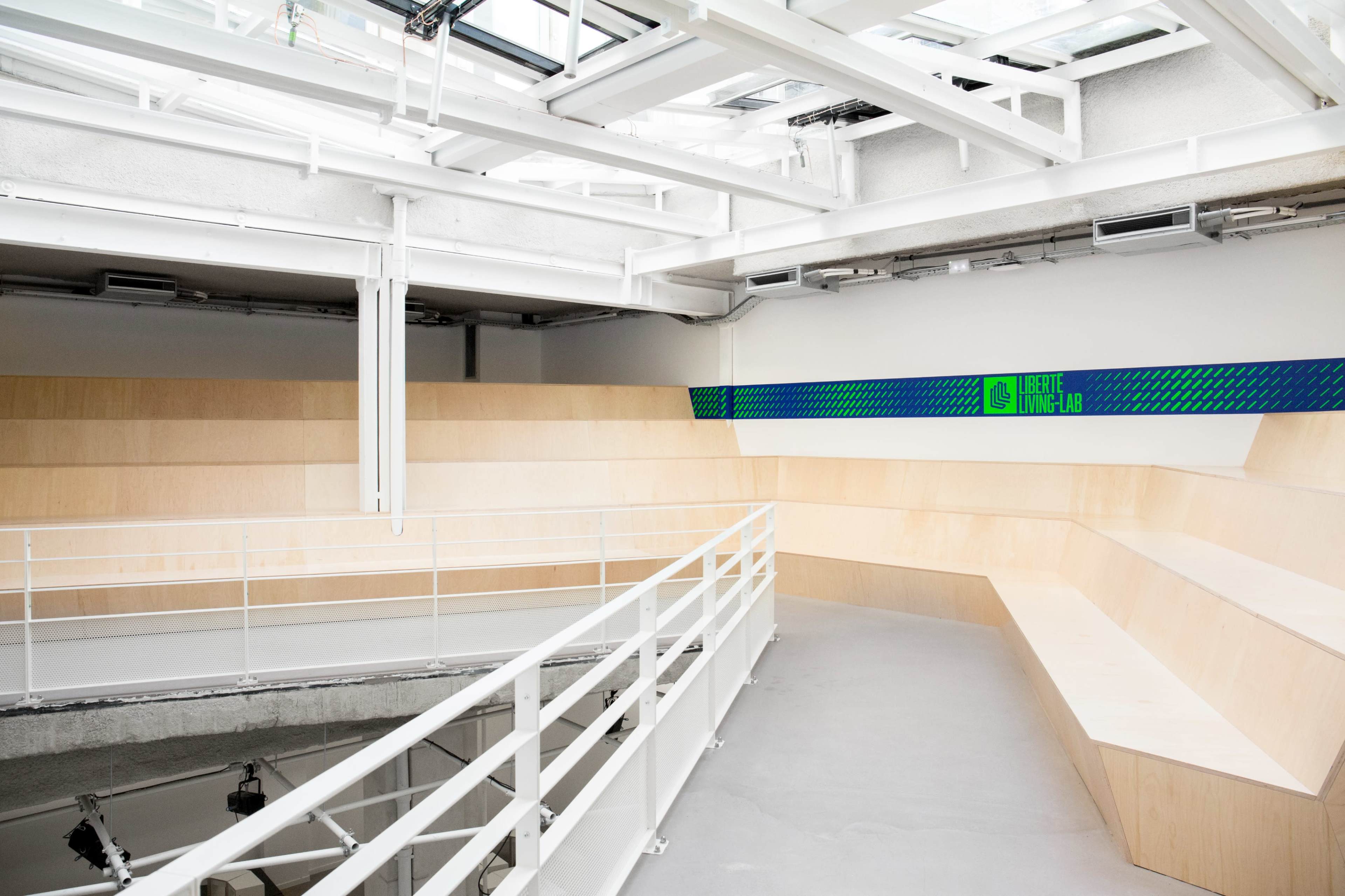 A modern, minimalist interior space featuring wooden seating arranged in a semicircular design, under a skylight with a bright green and blue accent wall.
