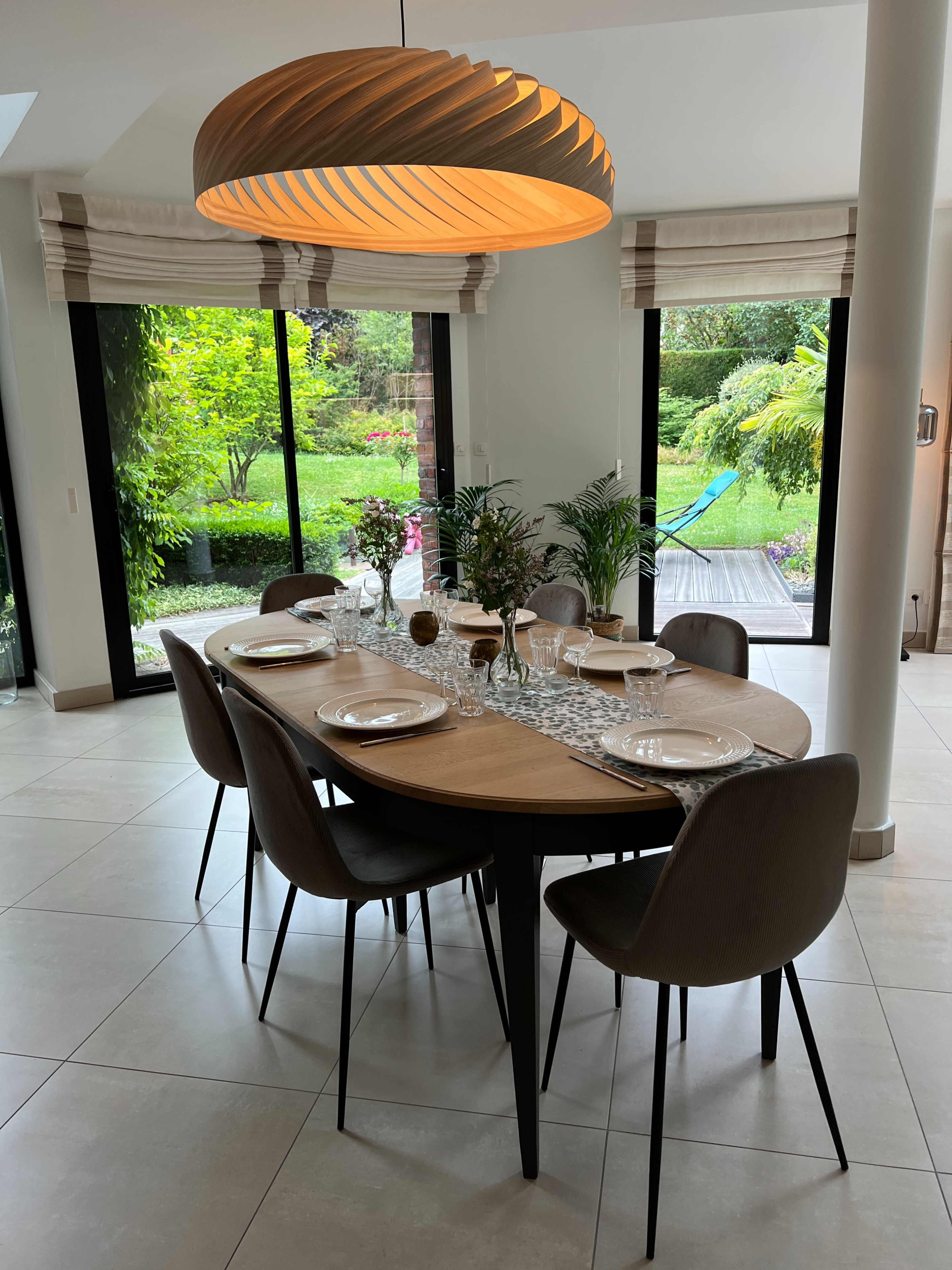 A dining table is set for a meal, featuring eight chairs, with a modern light fixture above and a view of a lush garden through large glass doors.