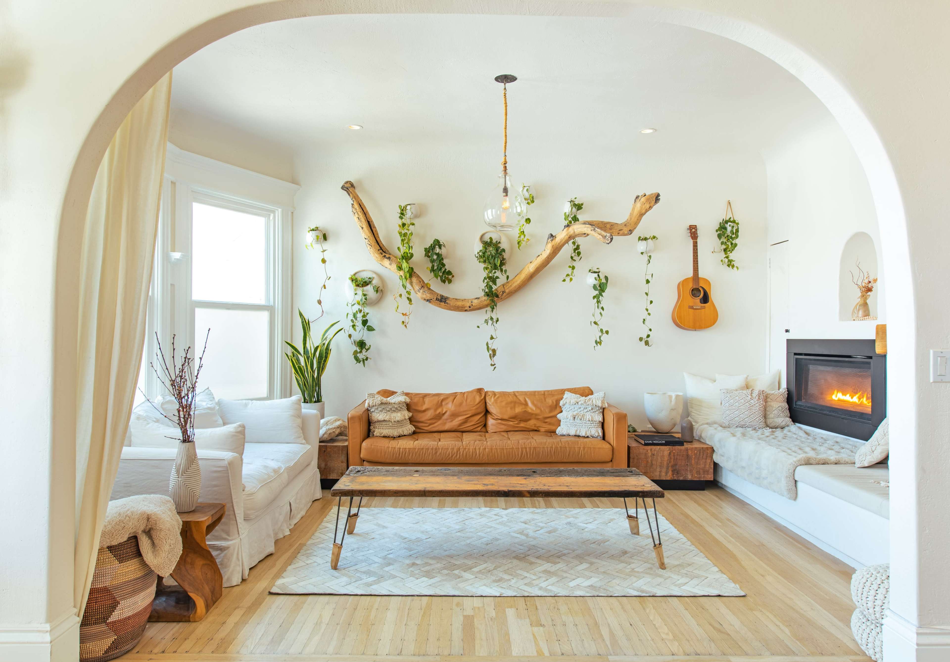 A bright living room features a leather sofa, a wooden coffee table, and plants hanging on the walls, with a guitar and a fireplace as focal points.