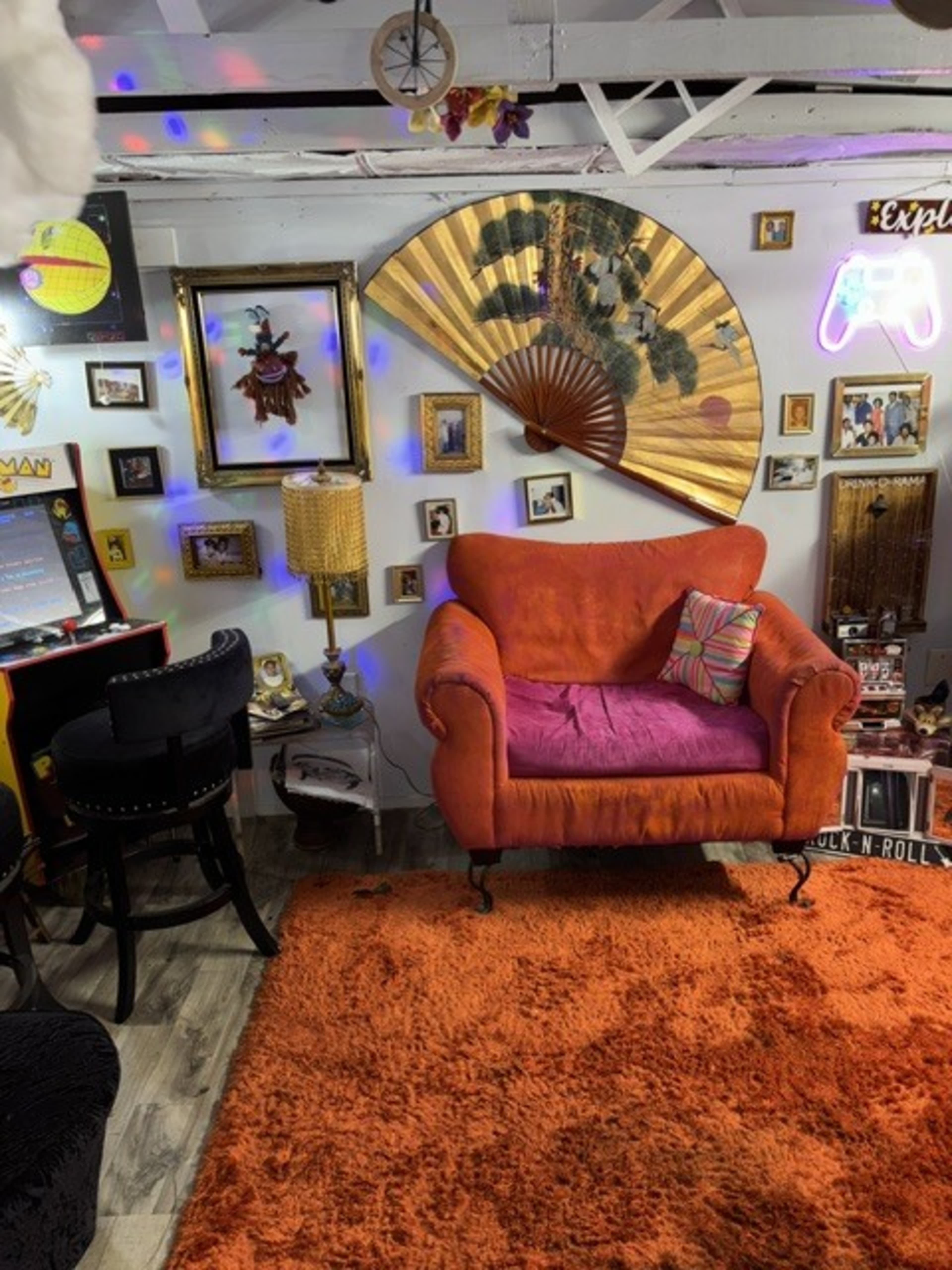 The image shows a colorful room featuring an orange upholstered chair, a large decorative fan on the wall, and various framed artworks and photographs arranged around the space.