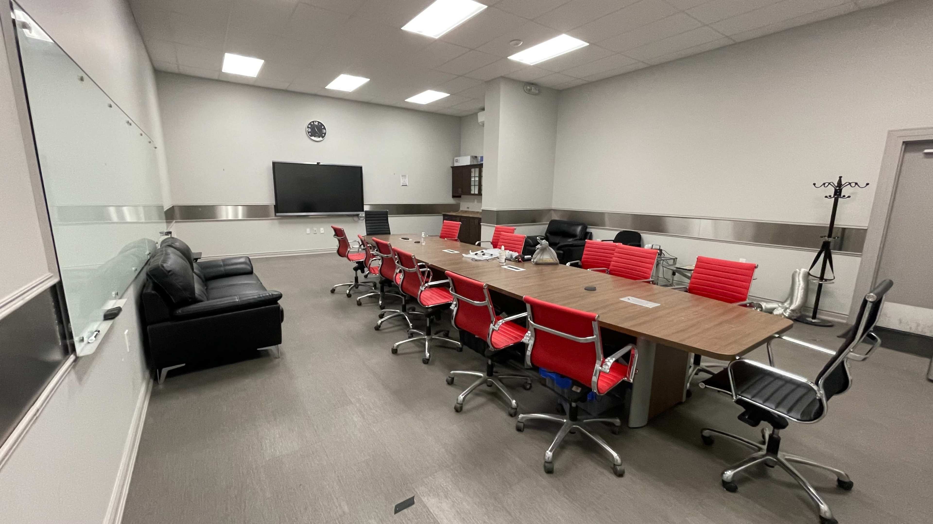A modern conference room features a long wooden table surrounded by red and black office chairs, with a flat-screen TV and a whiteboard on the walls.