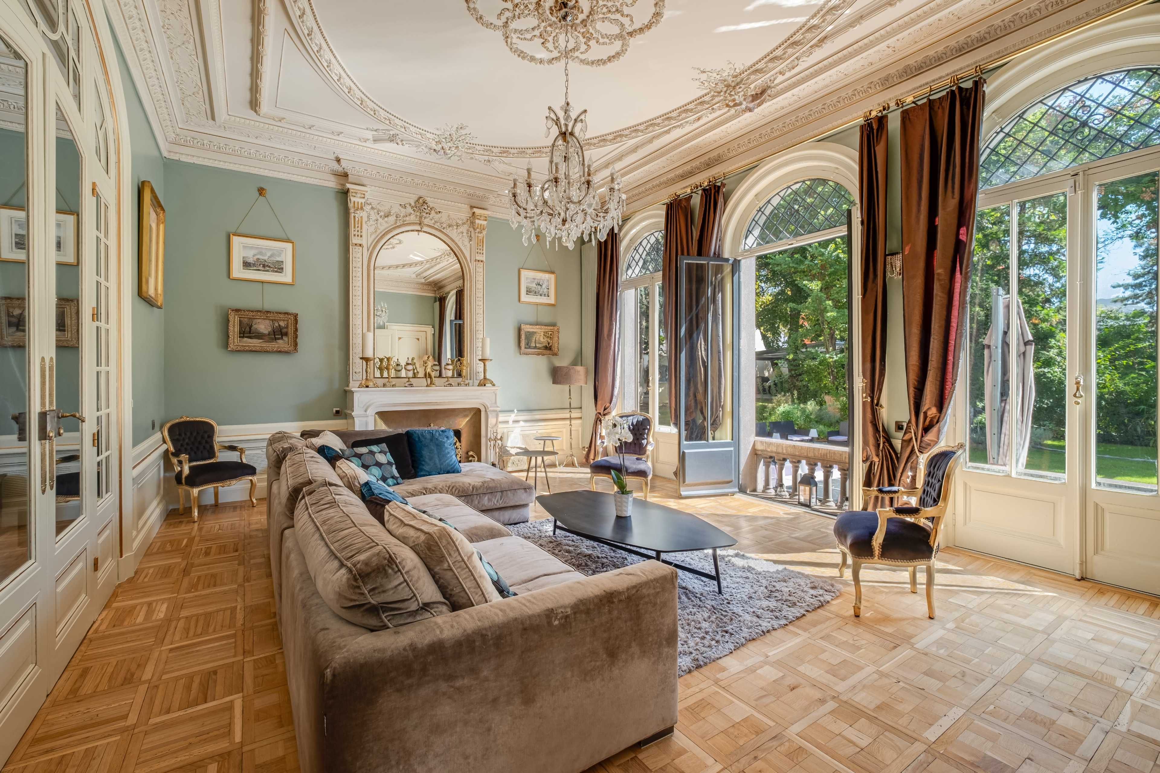 A spacious living room features a chandelier, large windows opening to a garden, and elegant furnishings arranged around a coffee table.