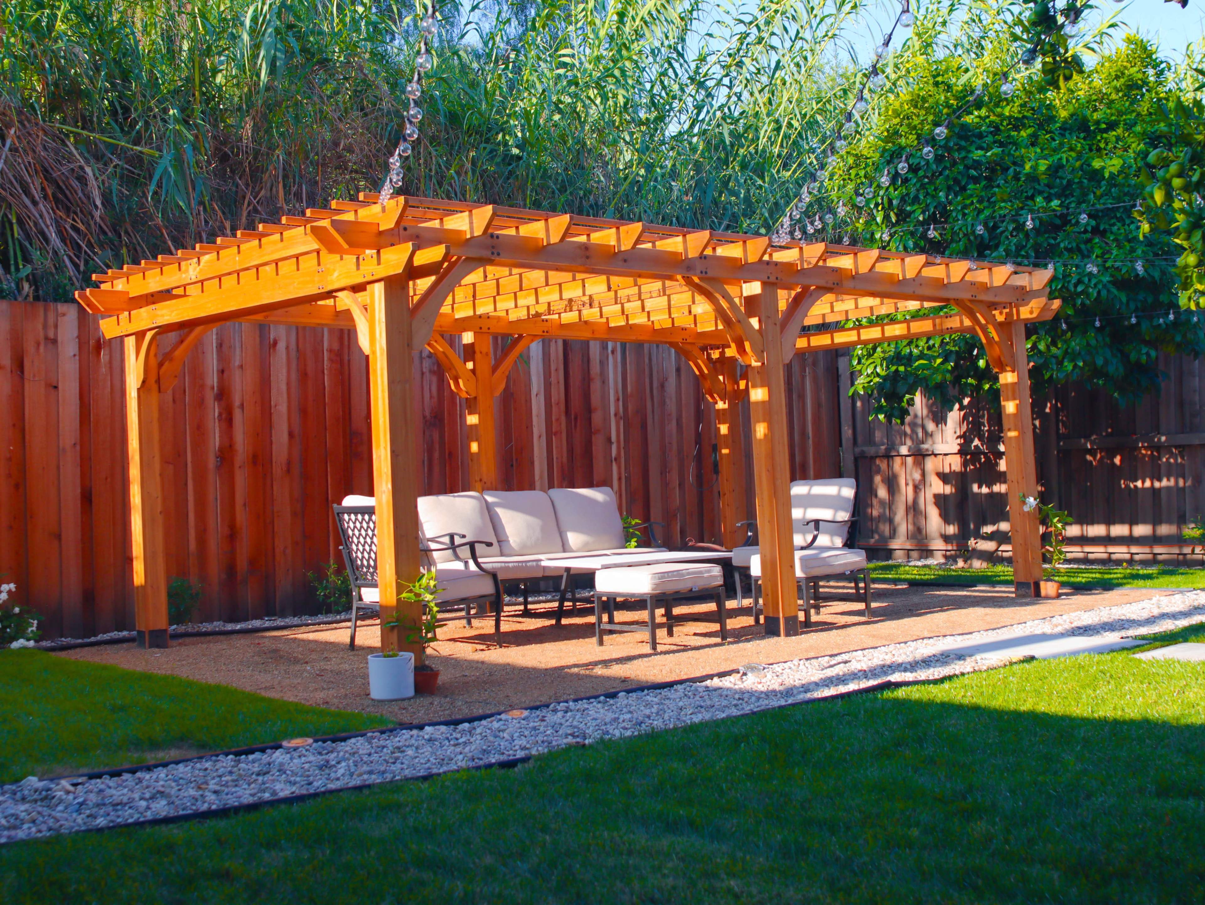 A wooden pergola furnished with seating on a grassy area surrounded by a wooden fence and vegetation.