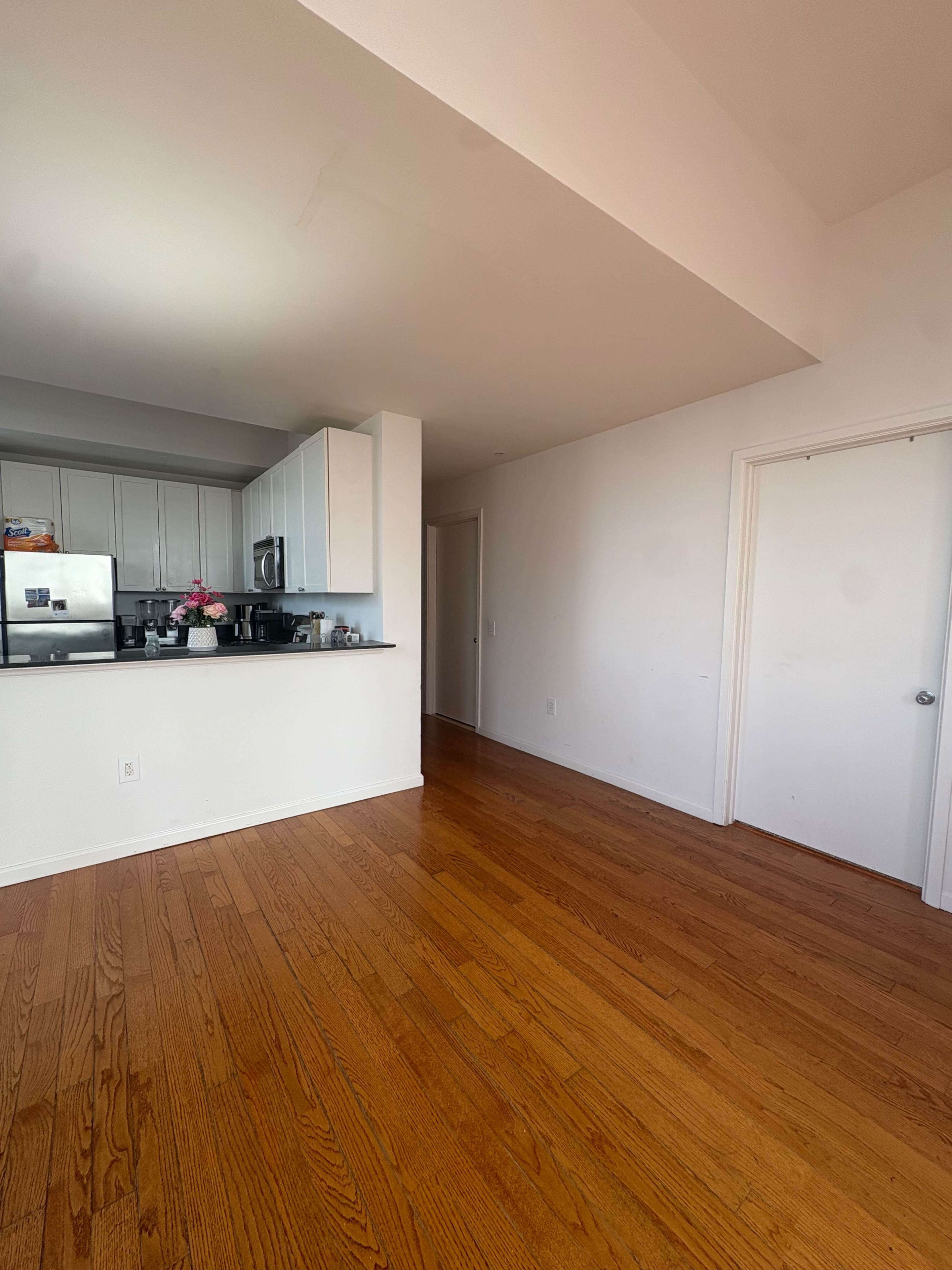 The image shows a spacious, well-lit room with hardwood floors, featuring a kitchen area with white cabinets and a door leading to another room.