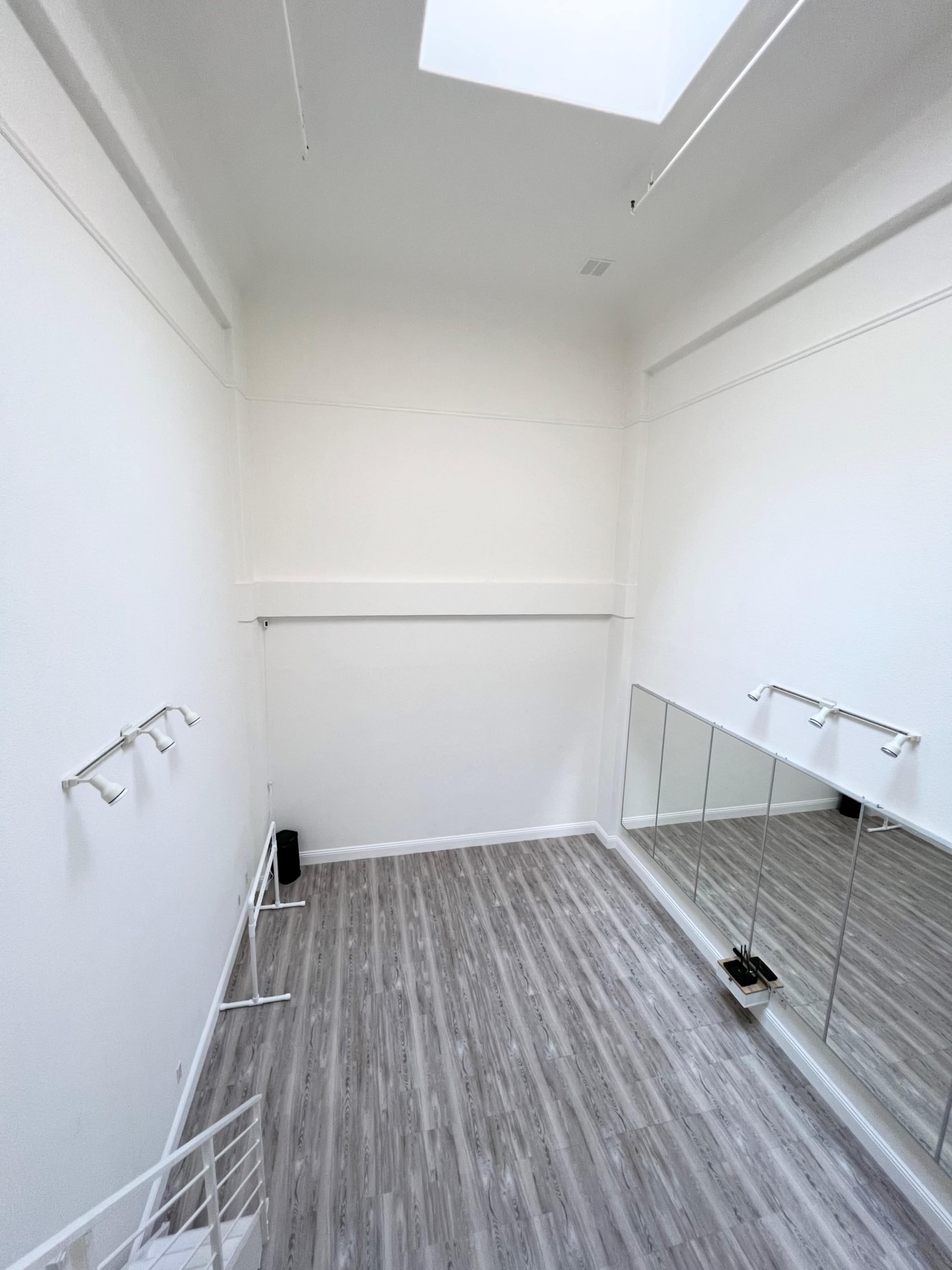A spacious, empty room with white walls, a skylight, and a mirrored wall, featuring light wooden flooring.