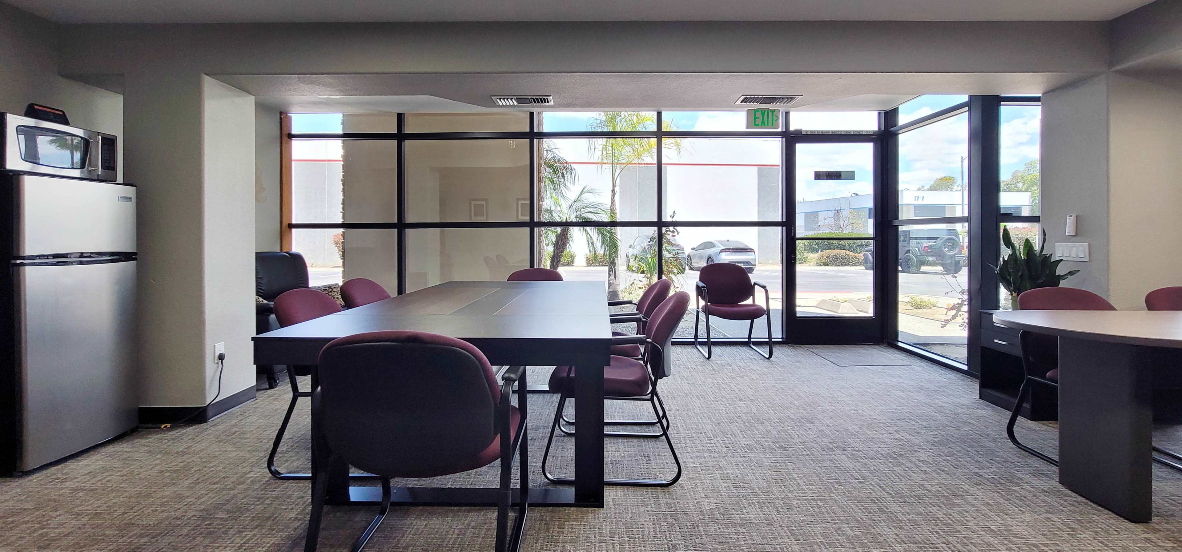The image shows a modern office space with a large table, several chairs, a refrigerator, and large windows that provide natural light.