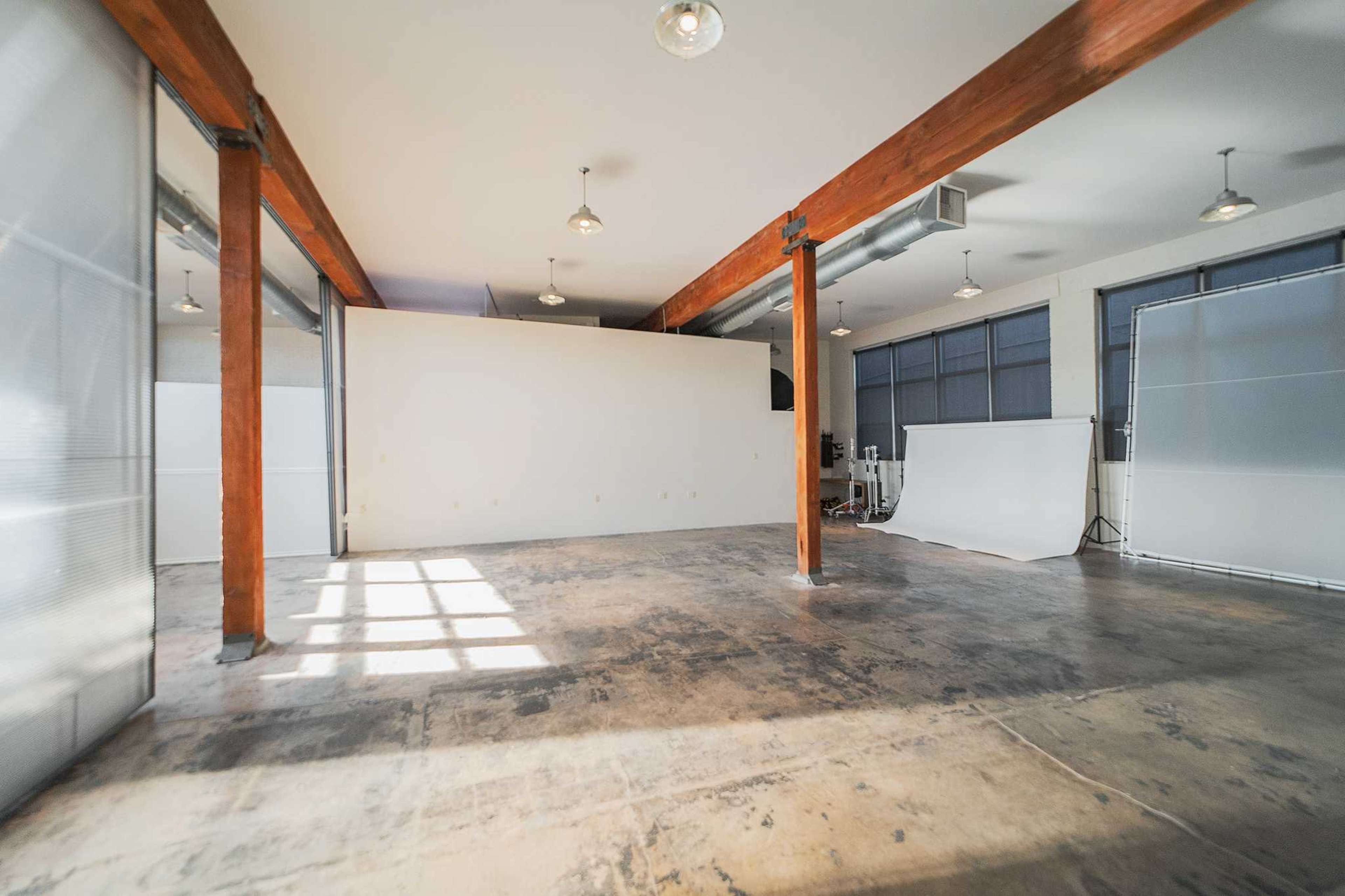 The image shows a spacious interior with exposed wooden beams, concrete flooring, and large windows that provide natural light.