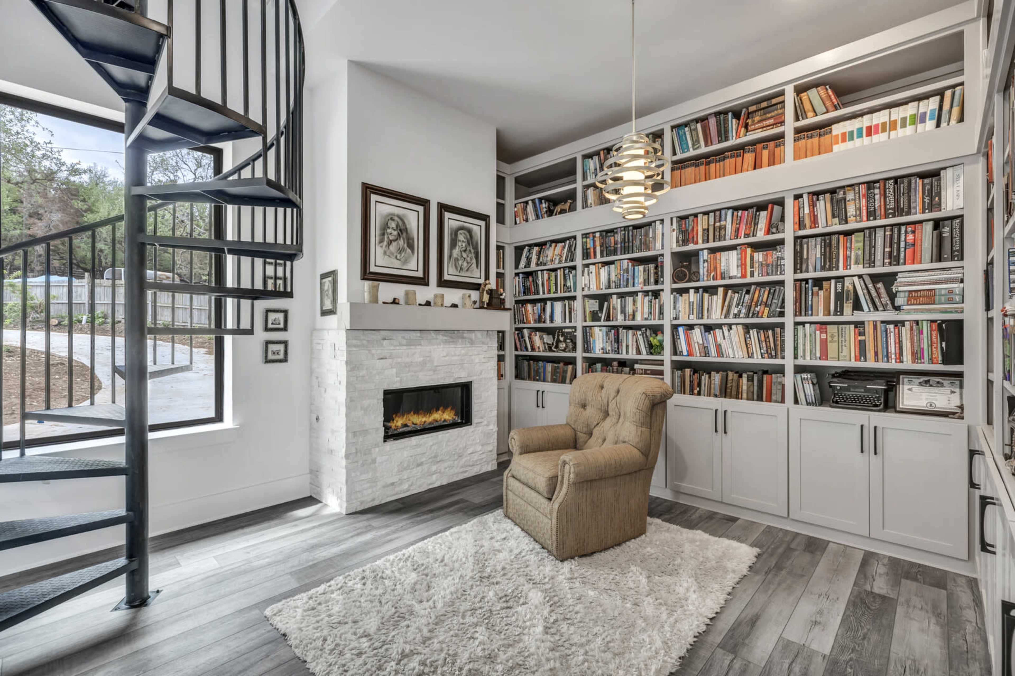 A cozy reading nook with a spiral staircase, a bookshelf filled with books, a plush armchair, and a modern fireplace against a white wall.