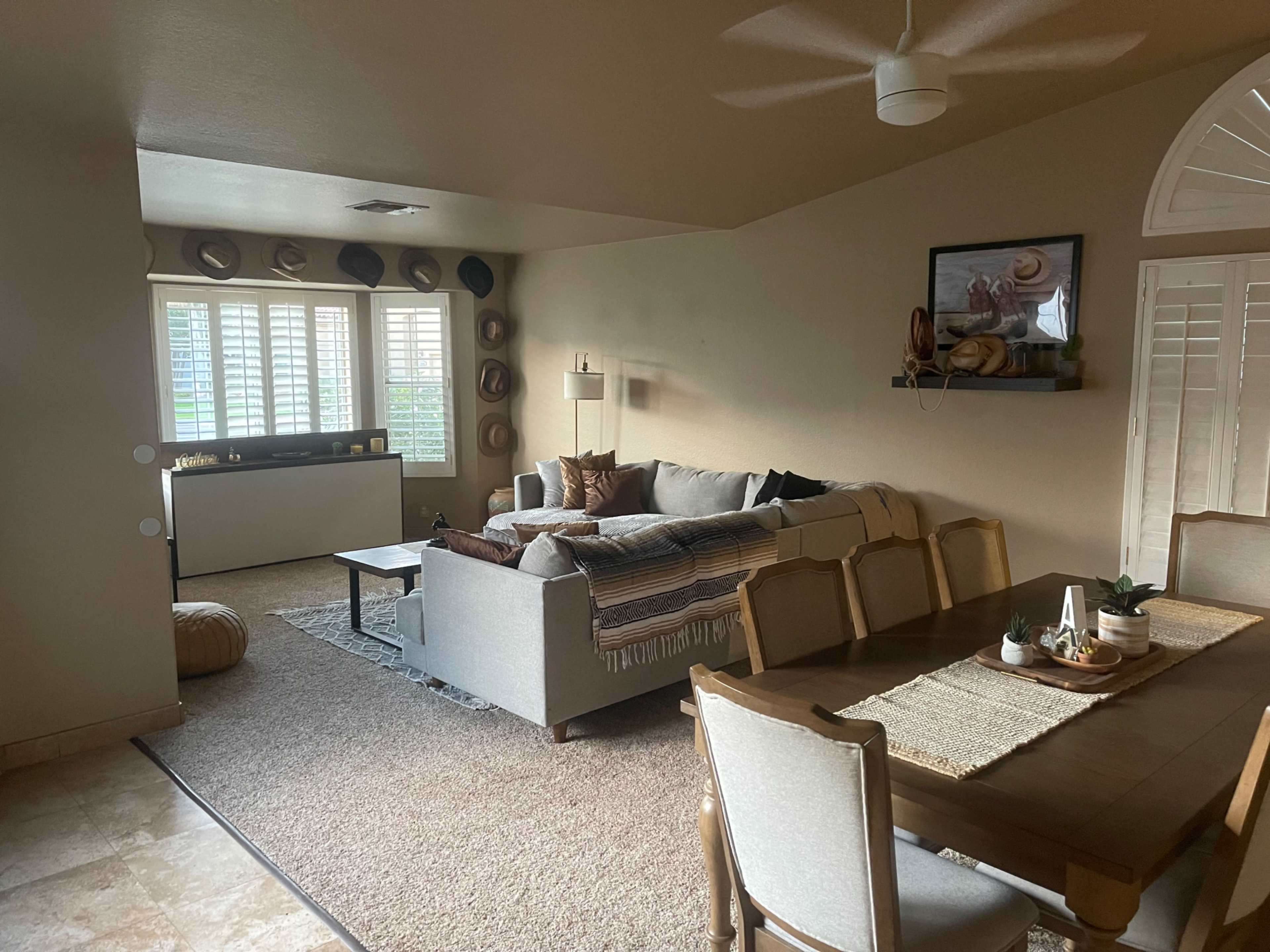 A living room features a gray sofa and a dining table with chairs, complemented by a light-colored area rug and decorative wall hangings.