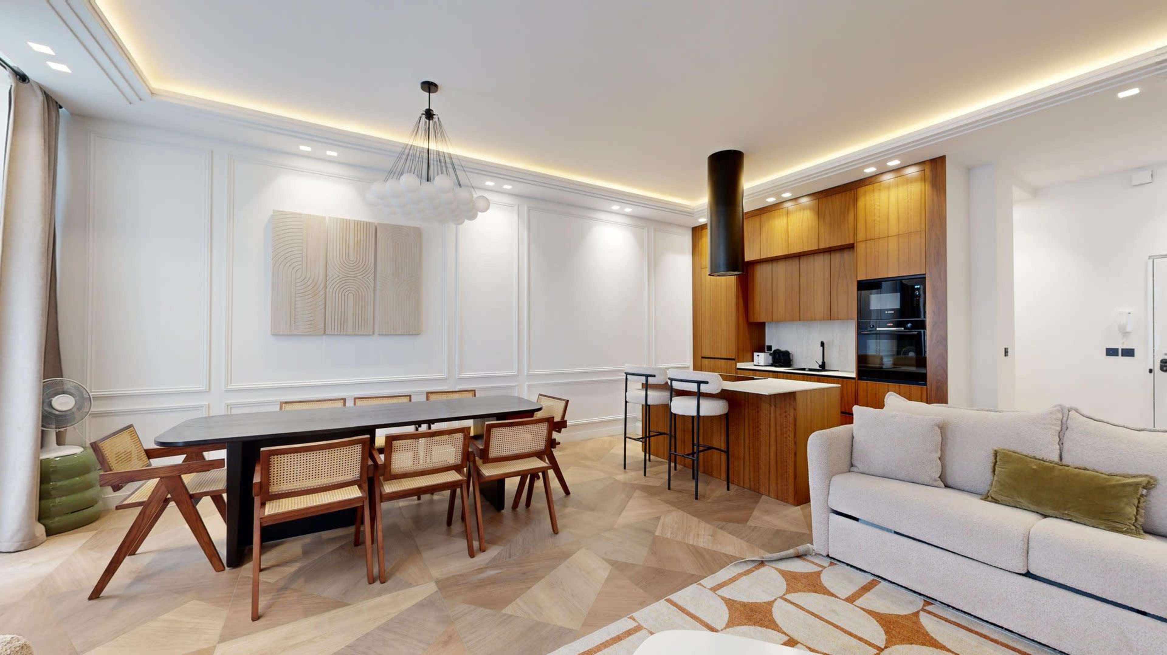The image shows a modern dining and kitchen area featuring a wooden dining table with chairs, a sleek kitchen with wooden cabinetry, and a cozy sofa in the living space.