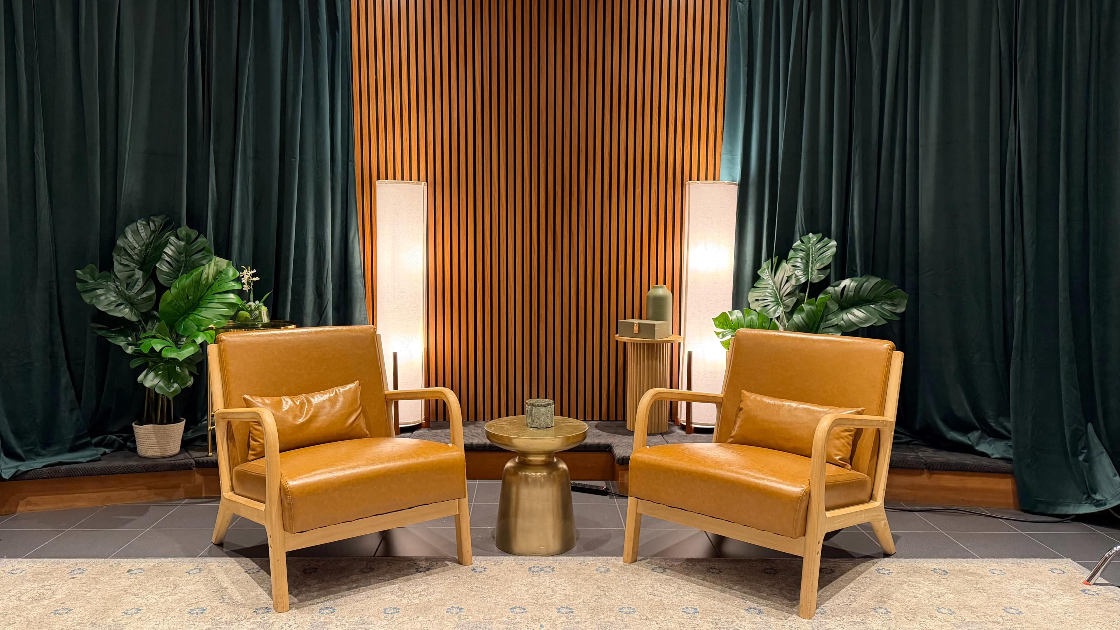 The image shows two orange armchairs placed on a patterned rug in front of a wooden slatted wall, flanked by plants and lit by floor lamps.