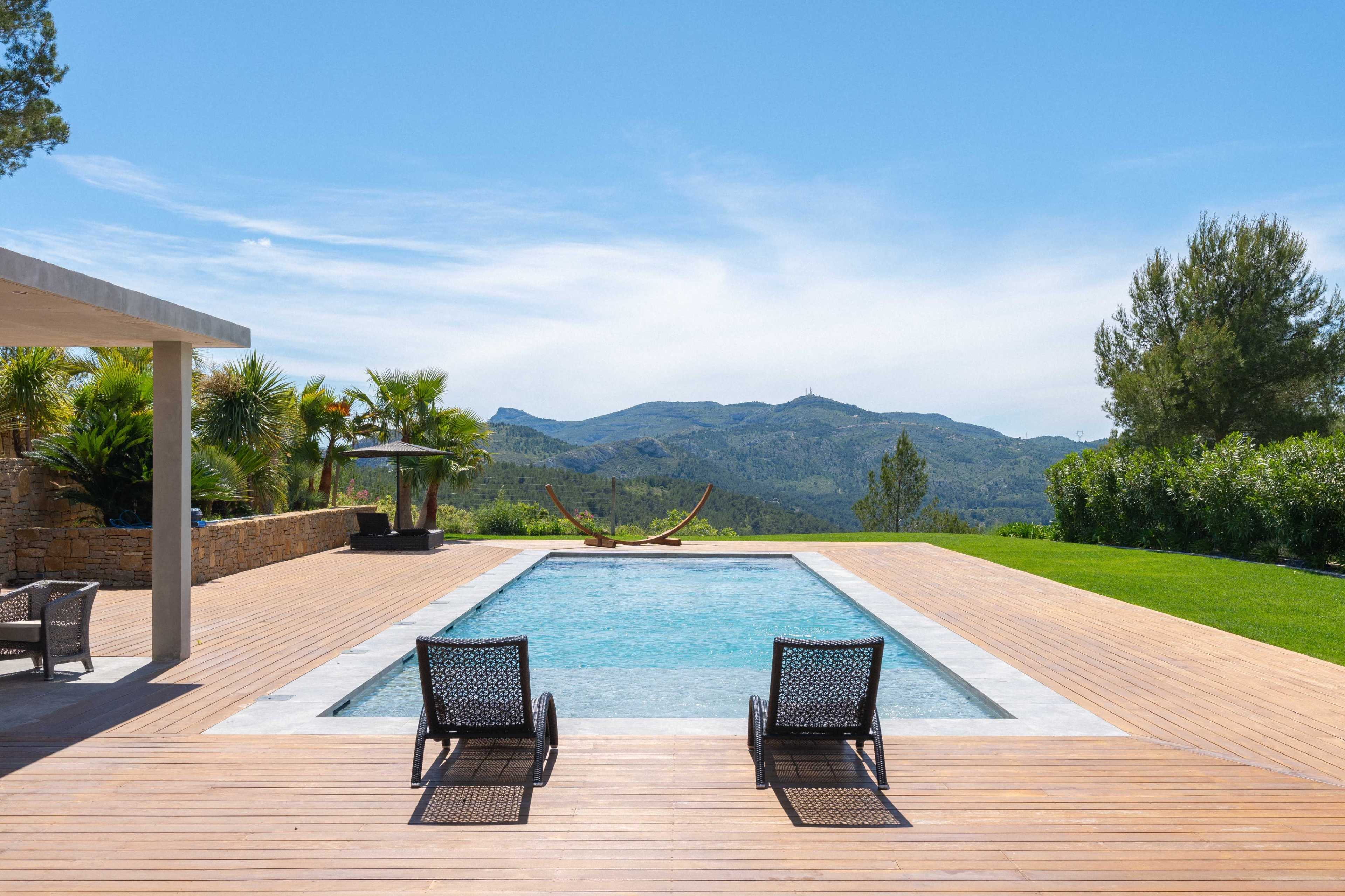 A modern pool surrounded by a wooden deck, with two lounge chairs facing the mountains in the background.