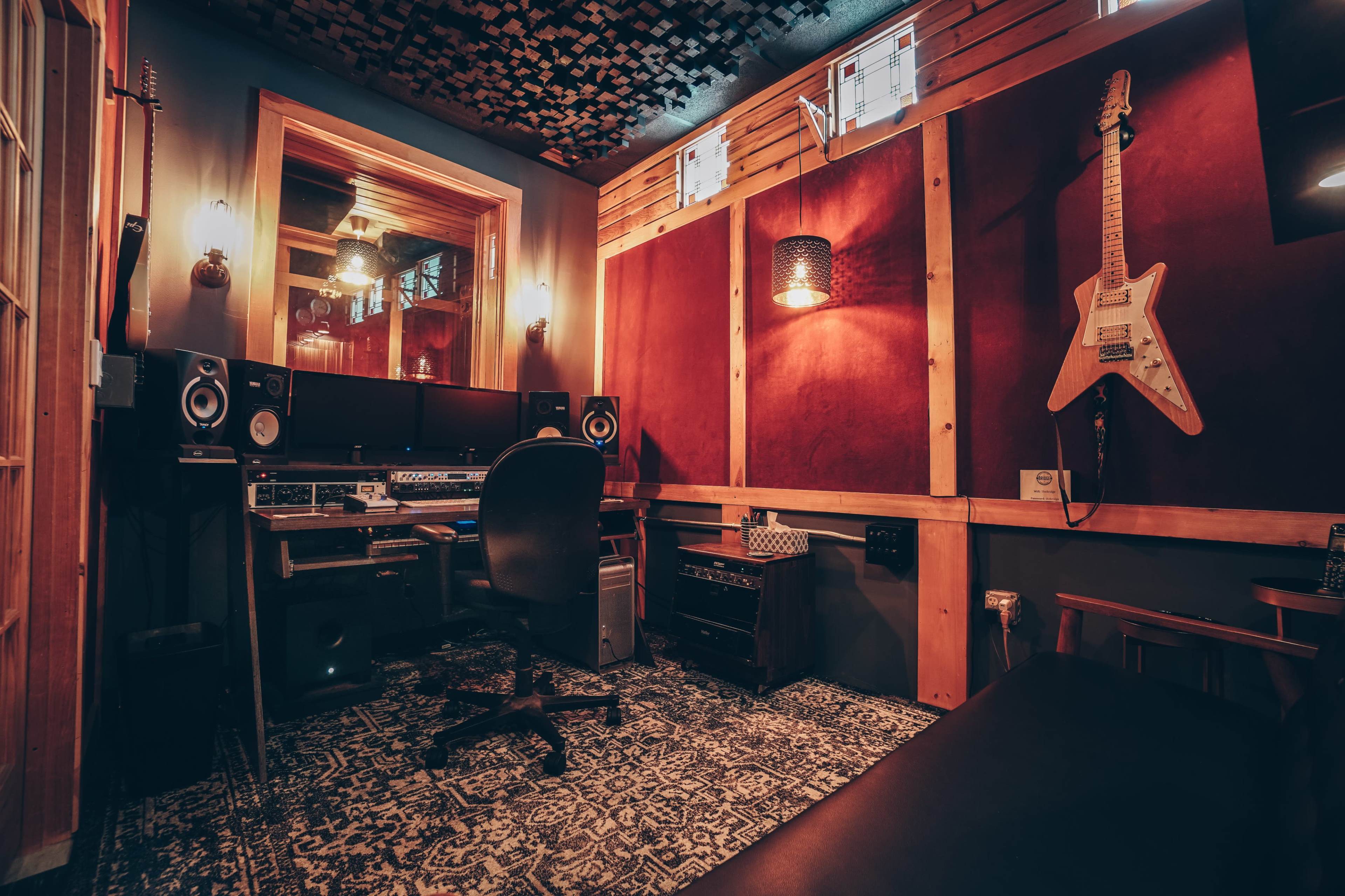 A recording studio with a large desk, computer equipment, speakers, and acoustic treatment on the walls, alongside a guitar mounted on a wooden panel.
