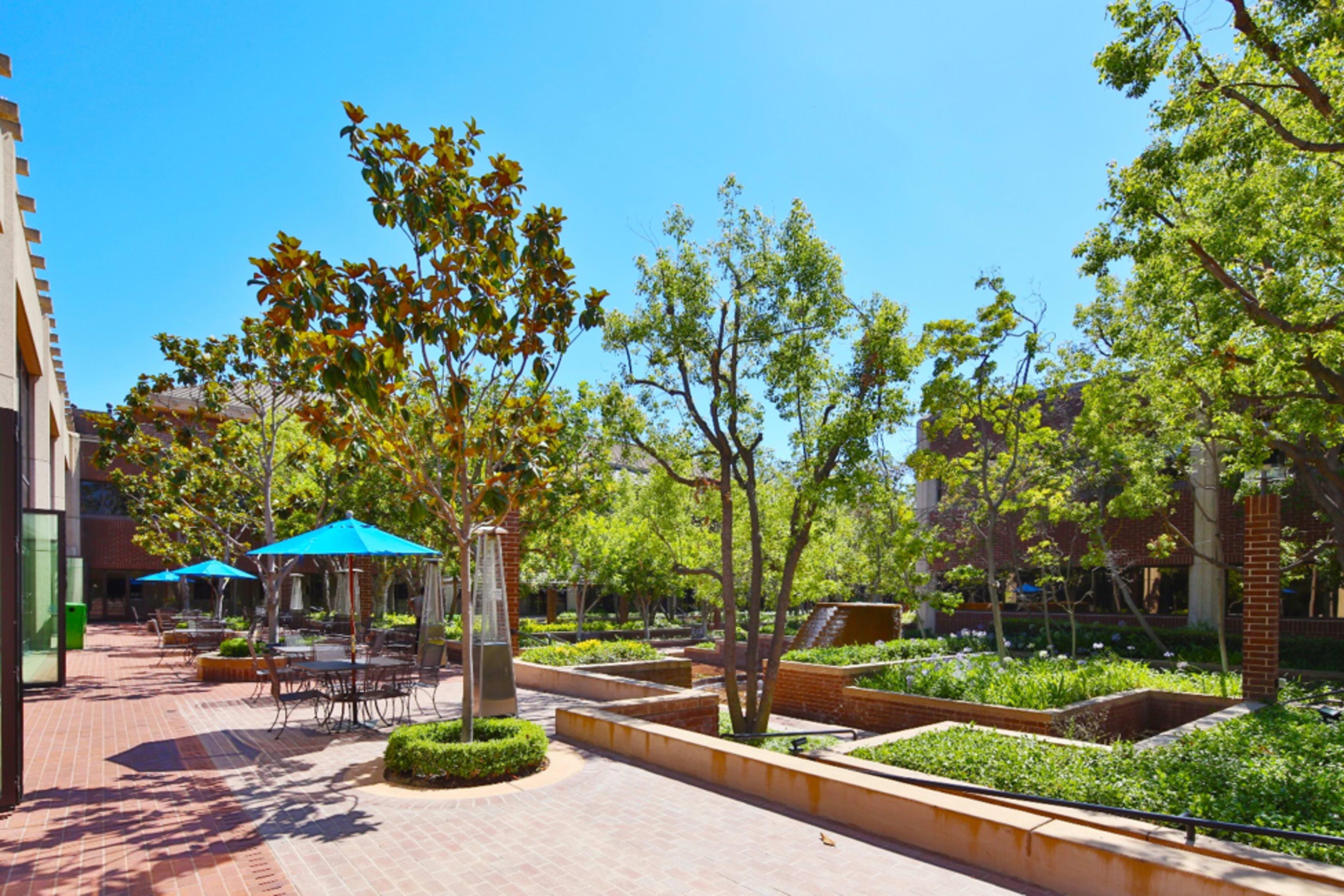 A landscaped courtyard features trees, seating areas with blue umbrellas, and rectangular garden beds.