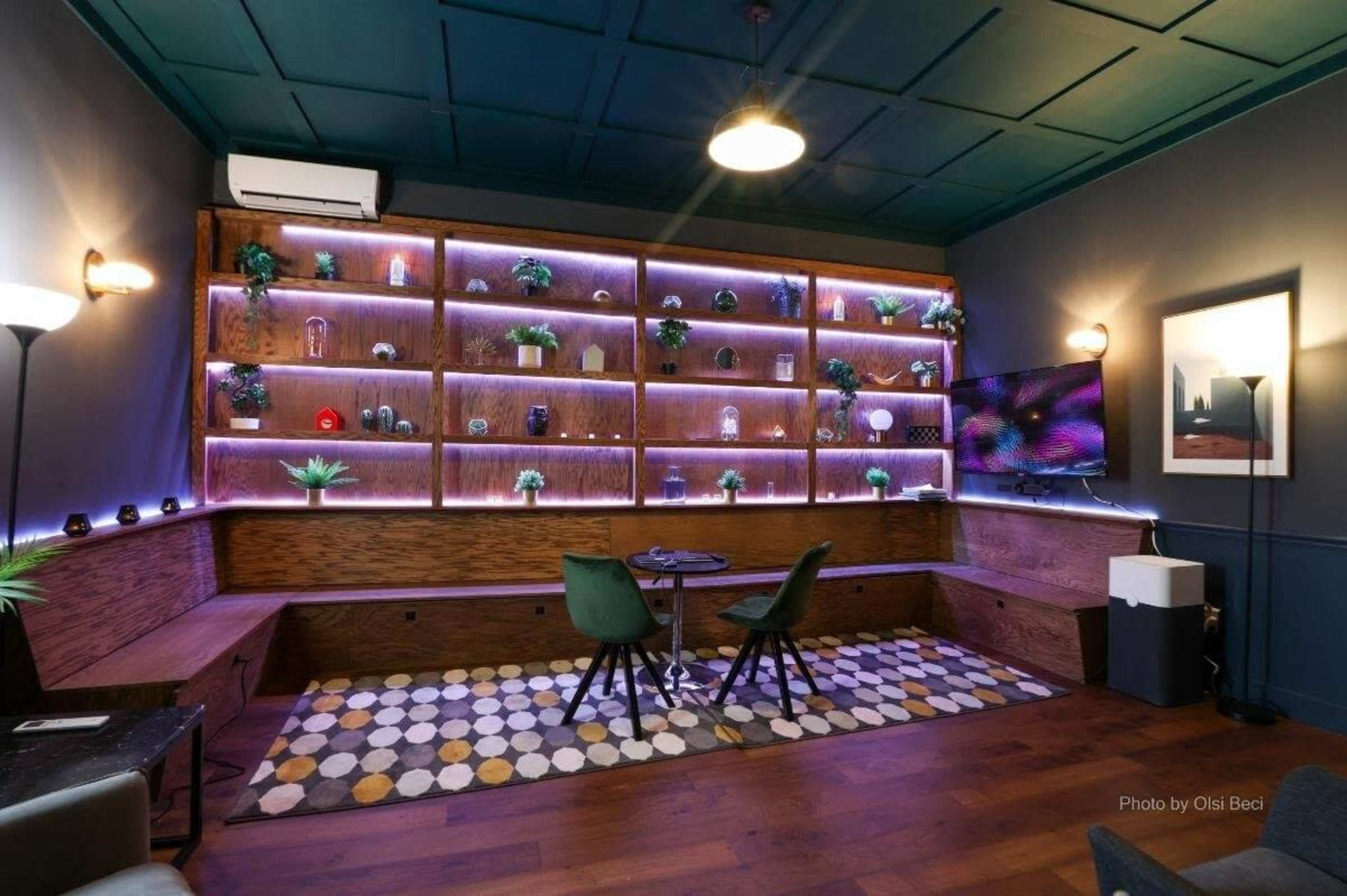 The image shows a modern interior space featuring wooden shelves with decorative items, a small sitting area with a round table and chairs, and ambient lighting along the shelves.