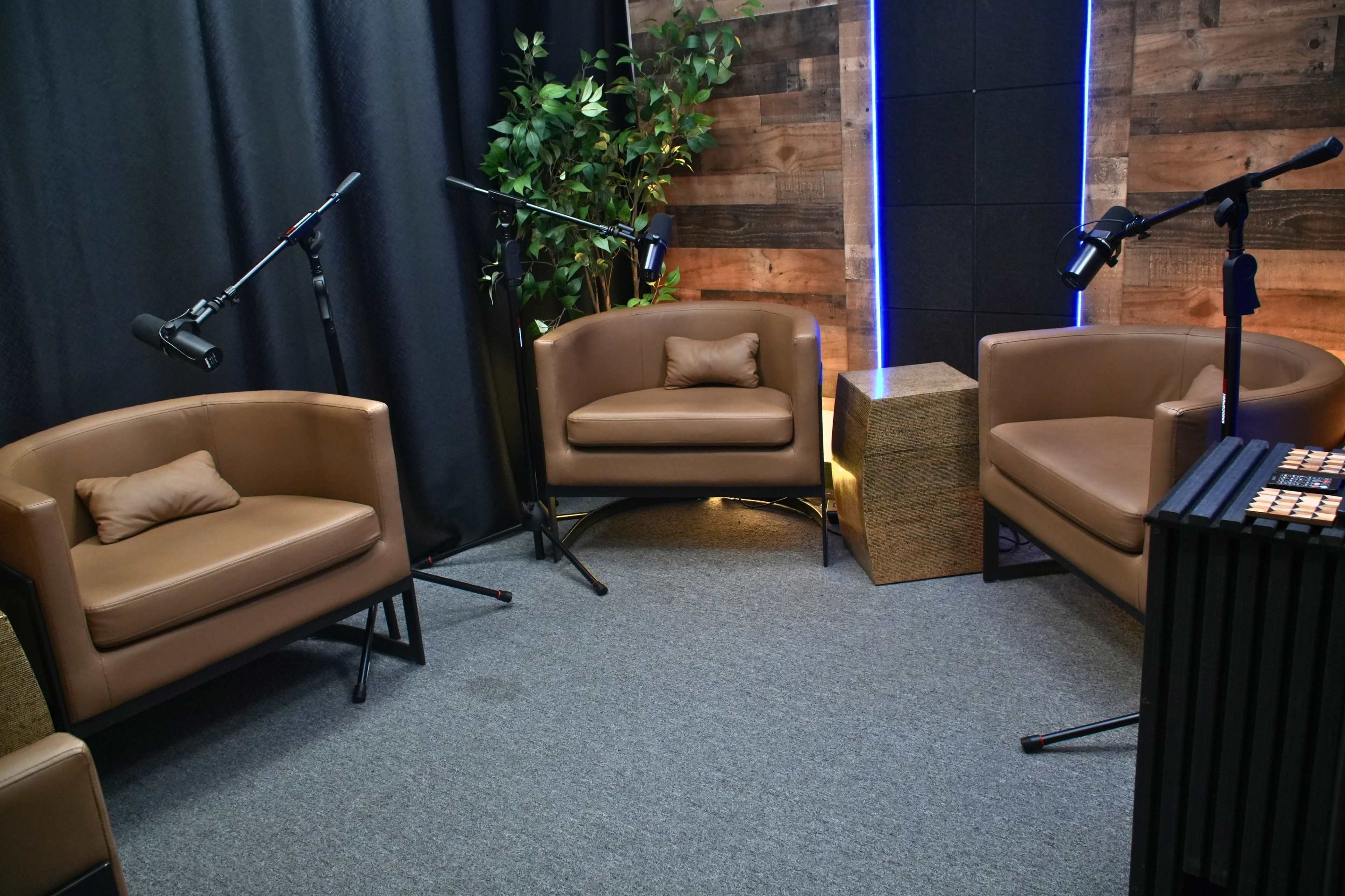 The image shows a small, modern podcast studio featuring two brown leather chairs, a decorative plant, and lighting accents against a wooden wall.