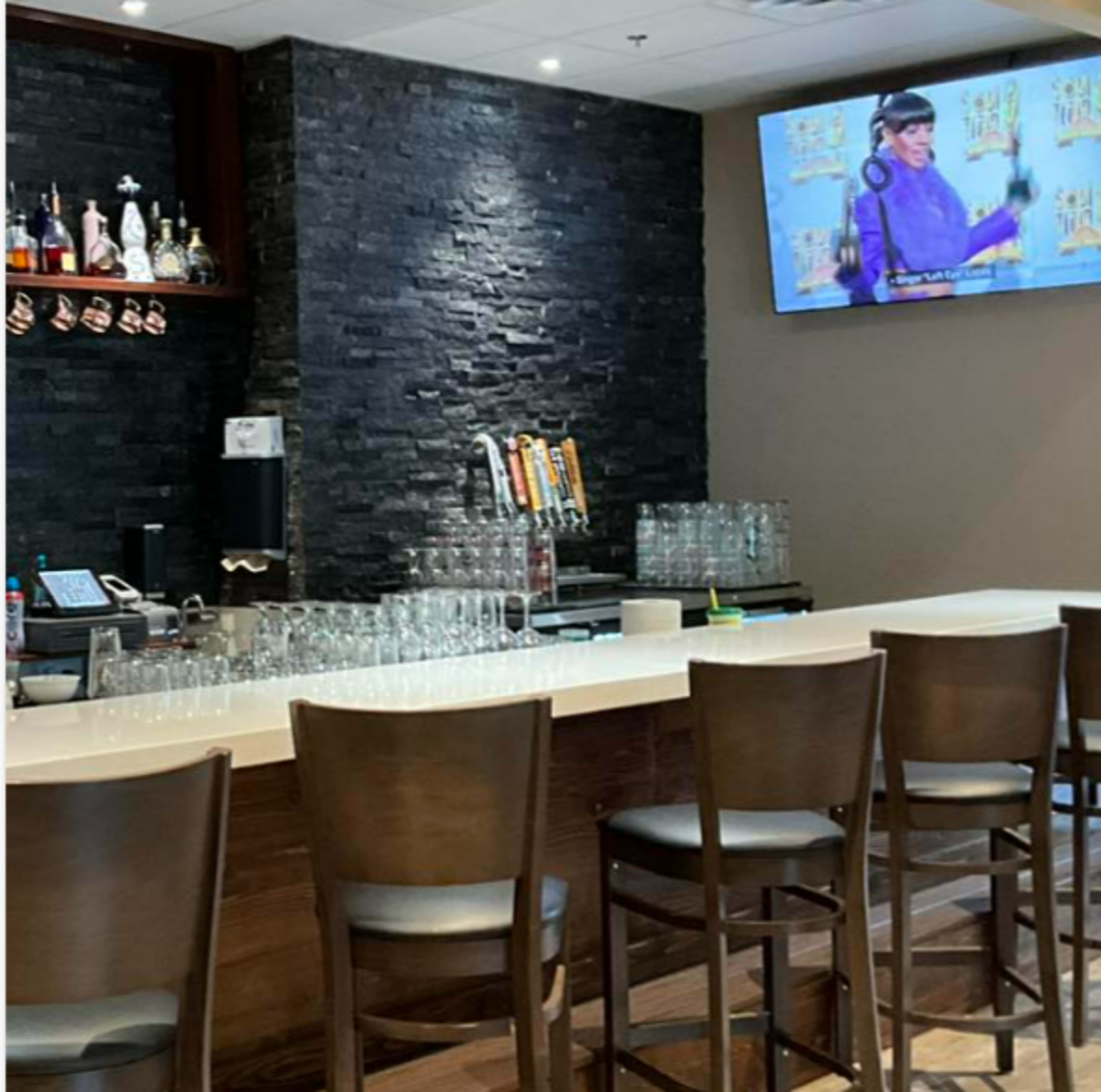 The image shows a modern bar area with a stone wall, a row of bar stools, and a television displaying a program.