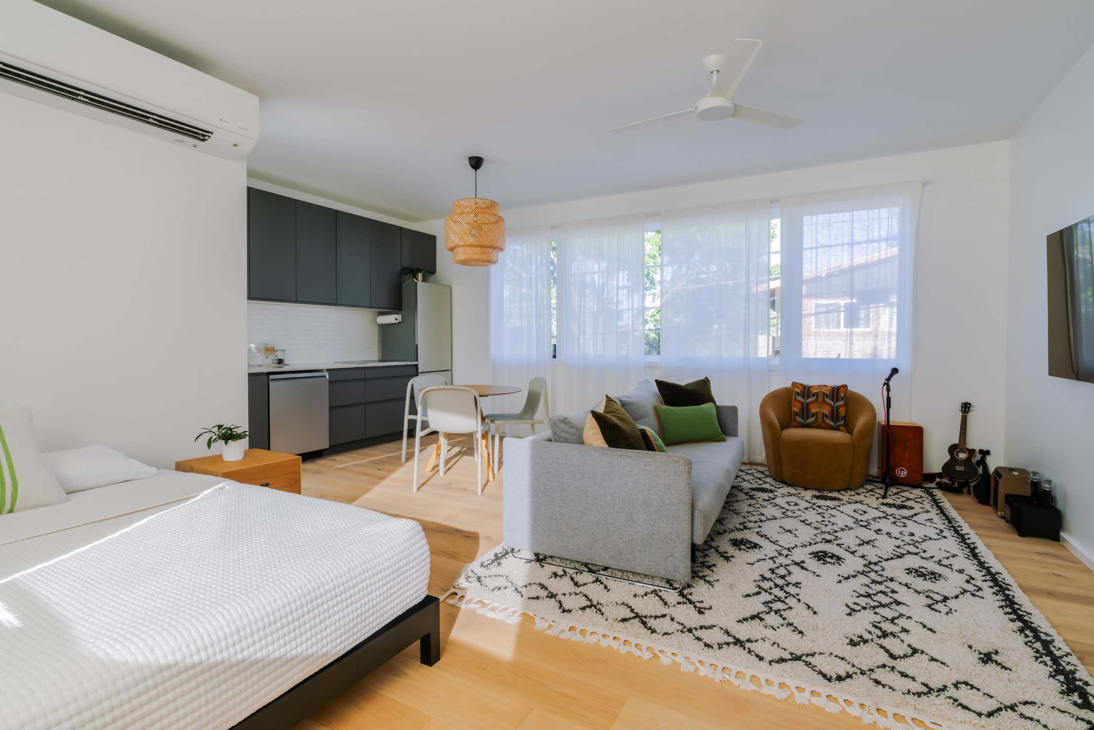 A modern living space features a gray sofa, wooden accents, a patterned rug, and a kitchen area in the background, illuminated by natural light through sheer curtains.