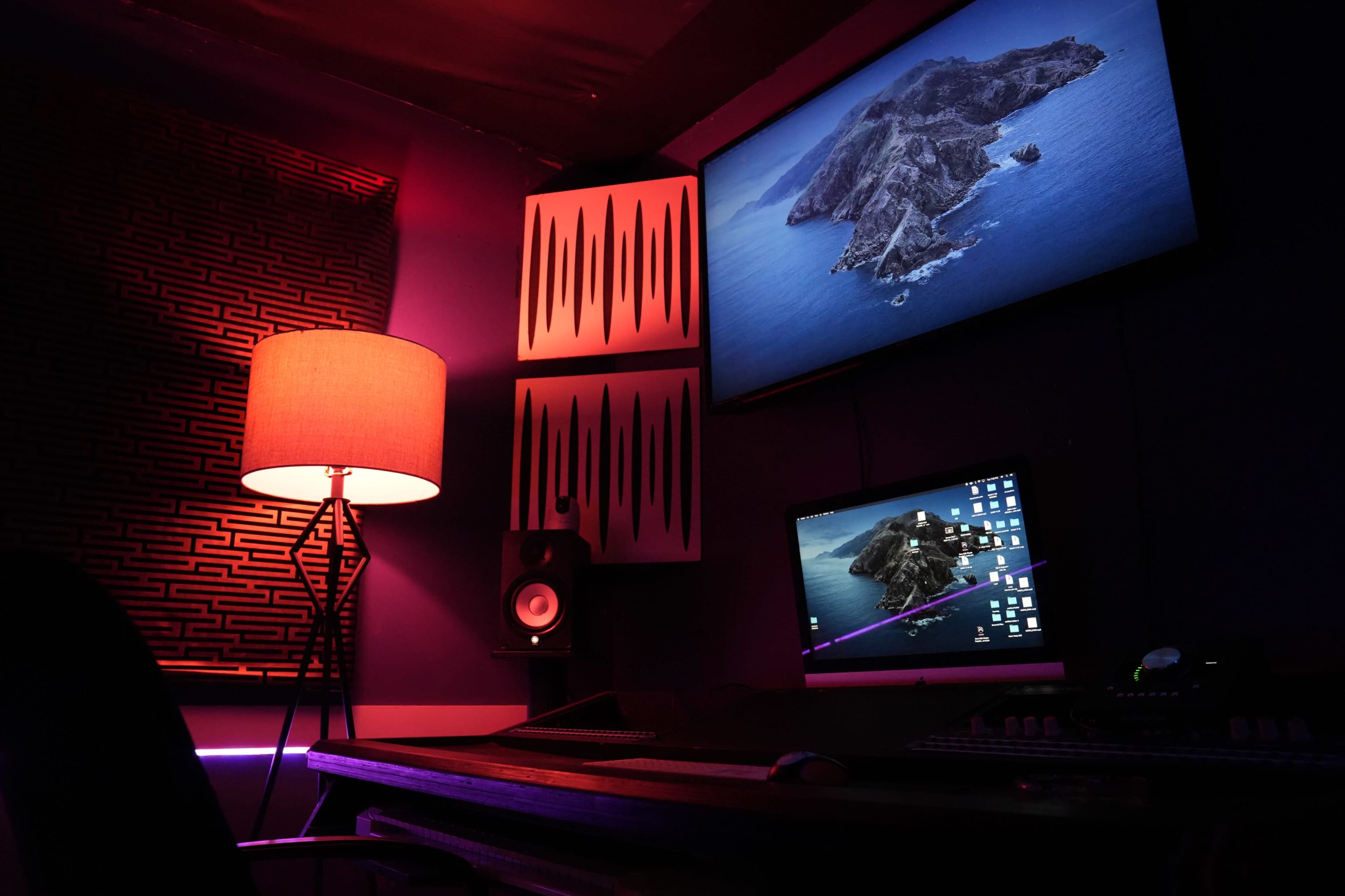 A dimly lit music studio features a desk with a computer and audio equipment, illuminated by a red lamp, and displays a scenic image of an island on the wall.