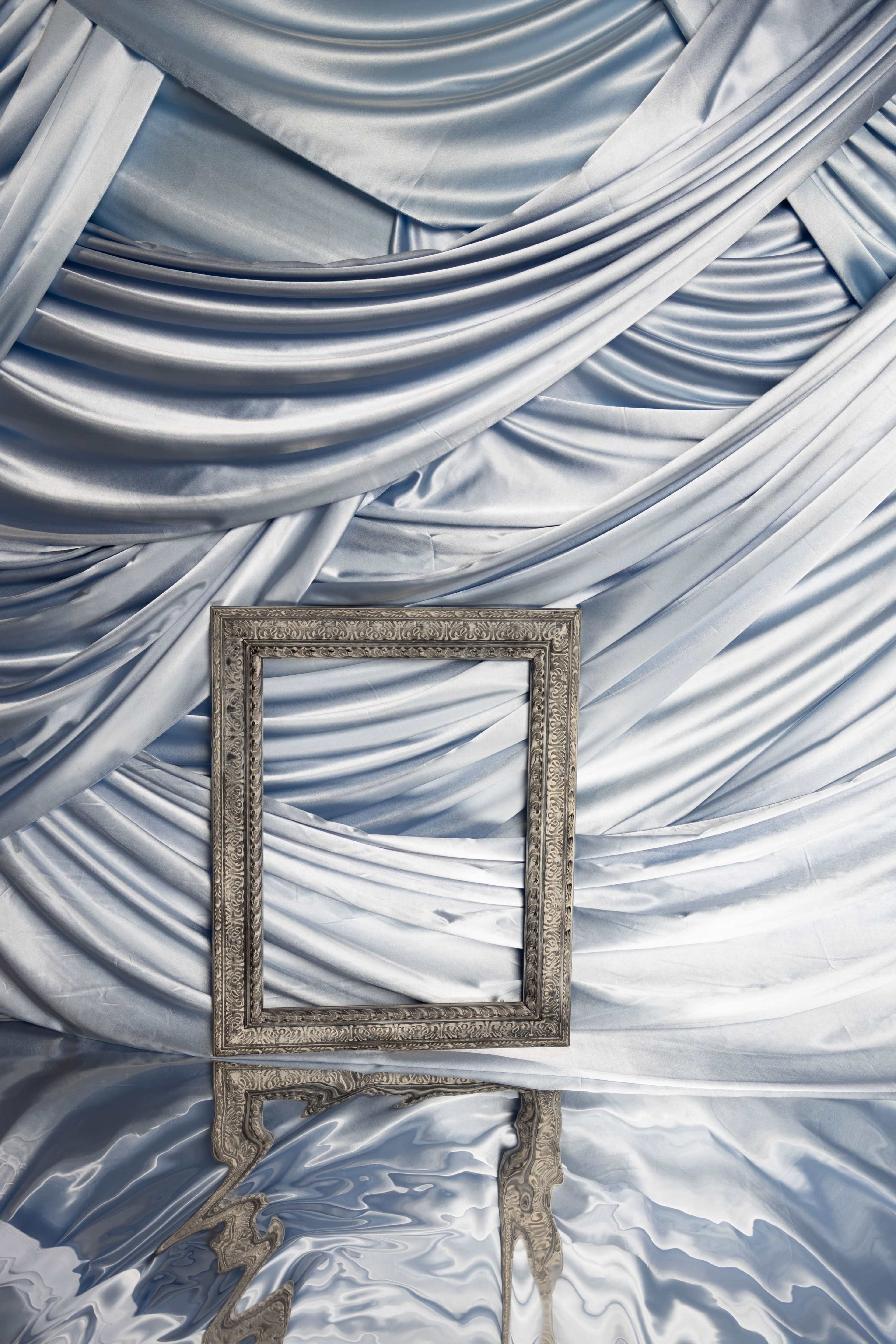 A silver ornate photo frame stands in front of a backdrop of draped blue fabric, reflecting in a smooth surface below.