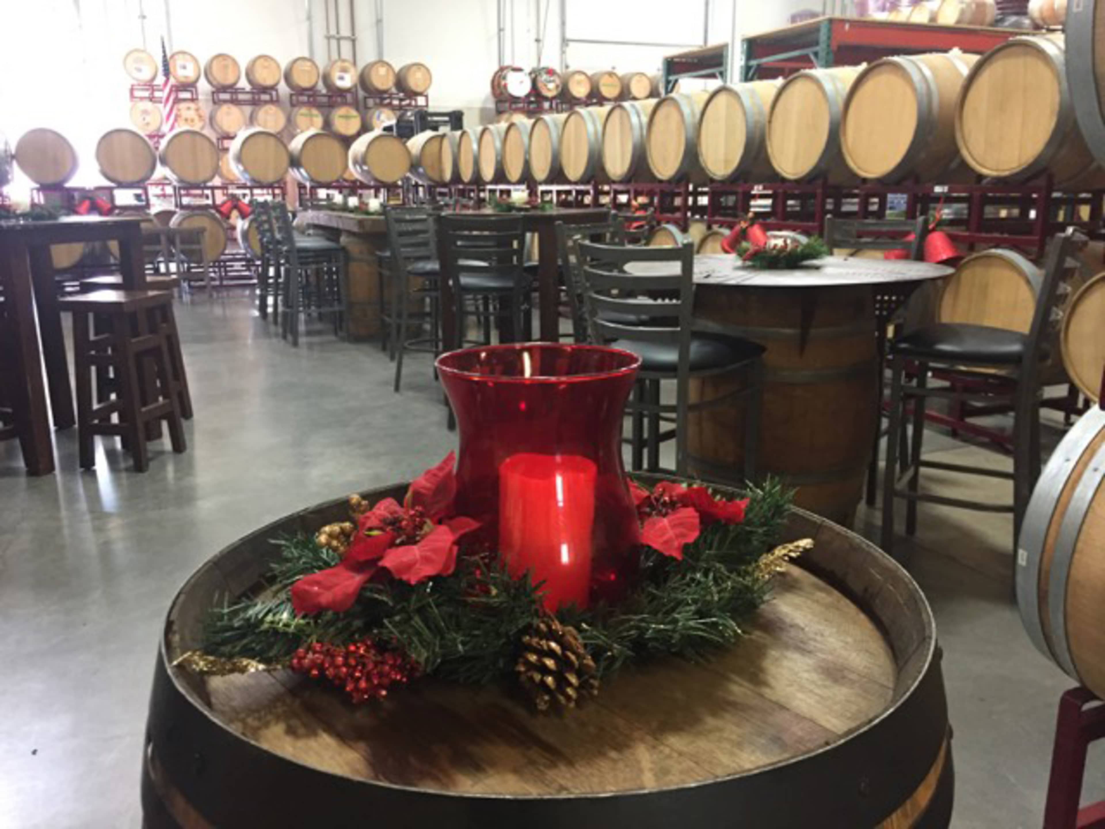A winery interior with wooden barrels, tables, and festive holiday decorations including a red candle surrounded by greenery.