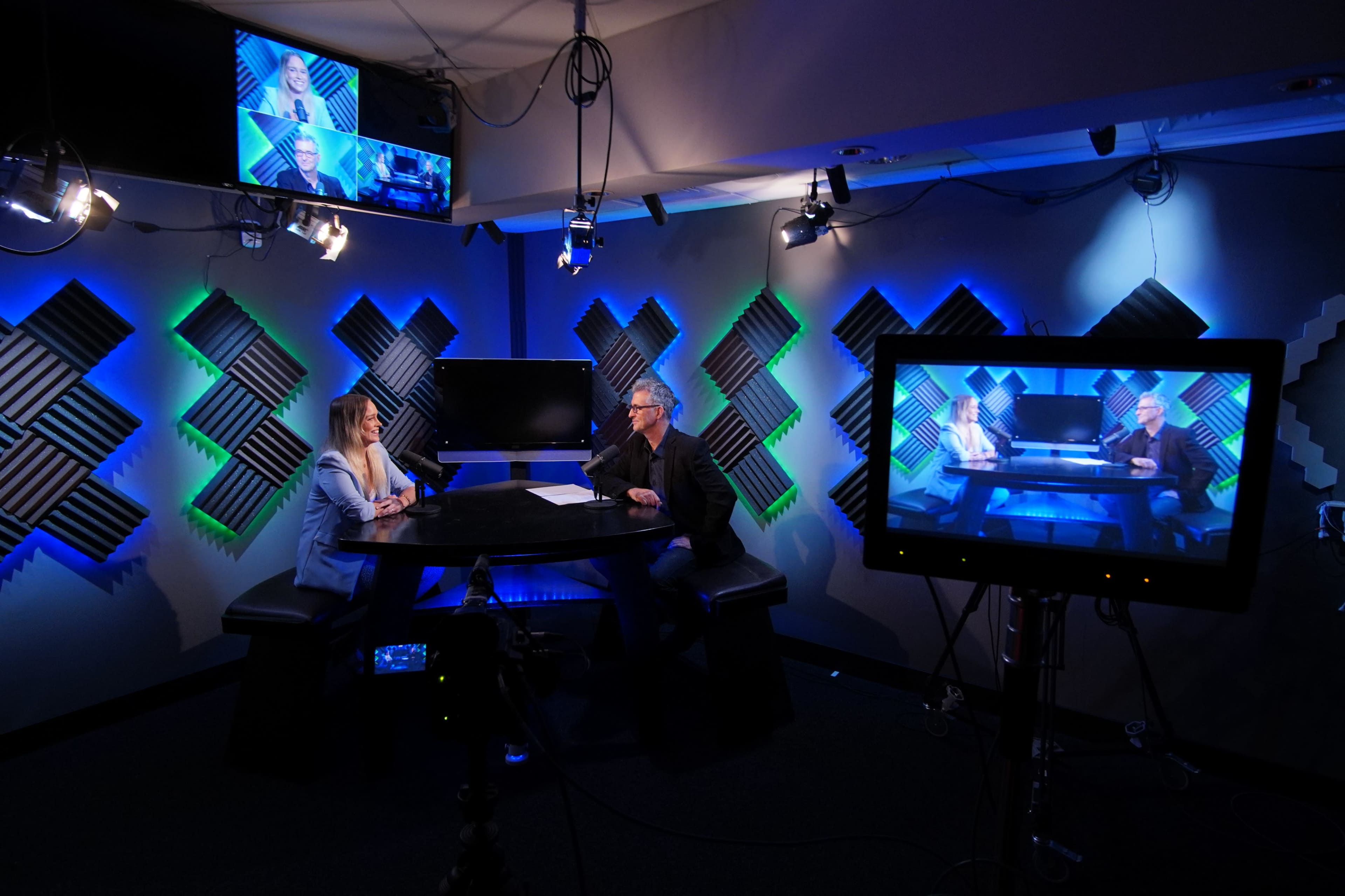 A television studio features two seated speakers at a black table, surrounded by blue and green lighting, with multiple screens displaying their discussion.