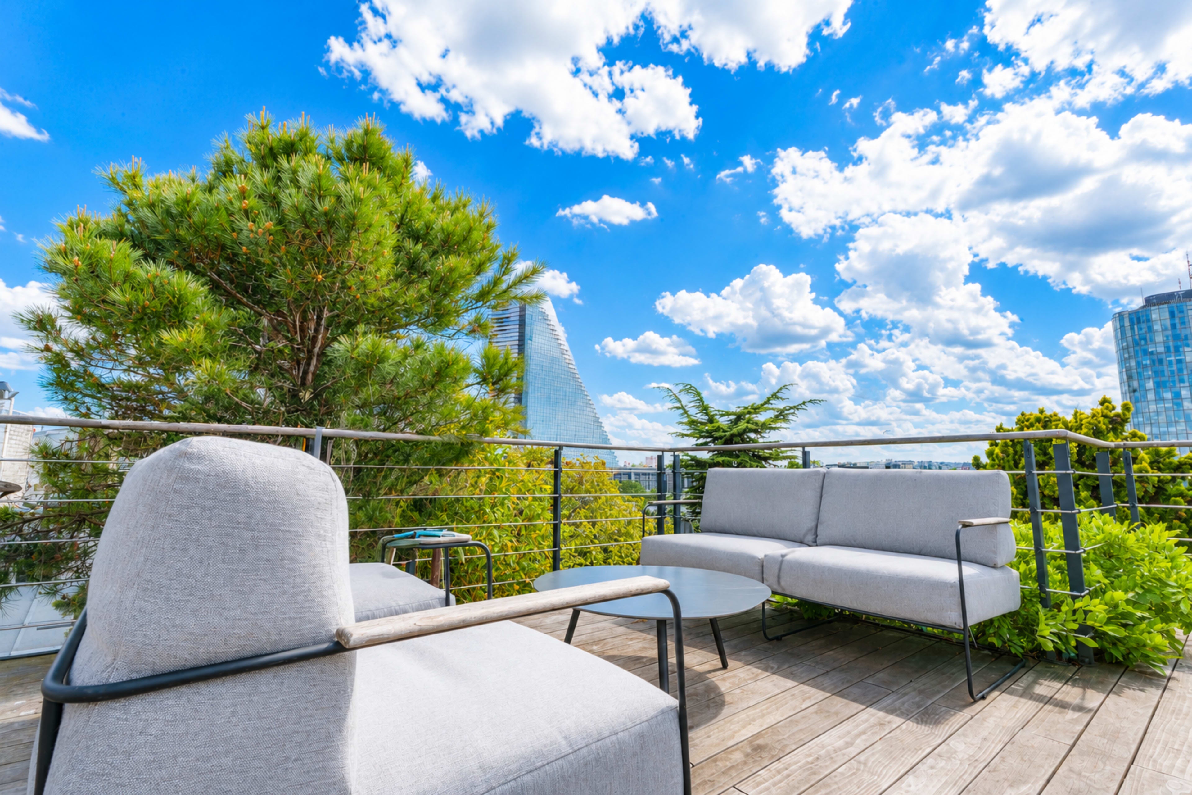 A rooftop terrace features modern seating arrangements surrounded by greenery under a partly cloudy sky.
