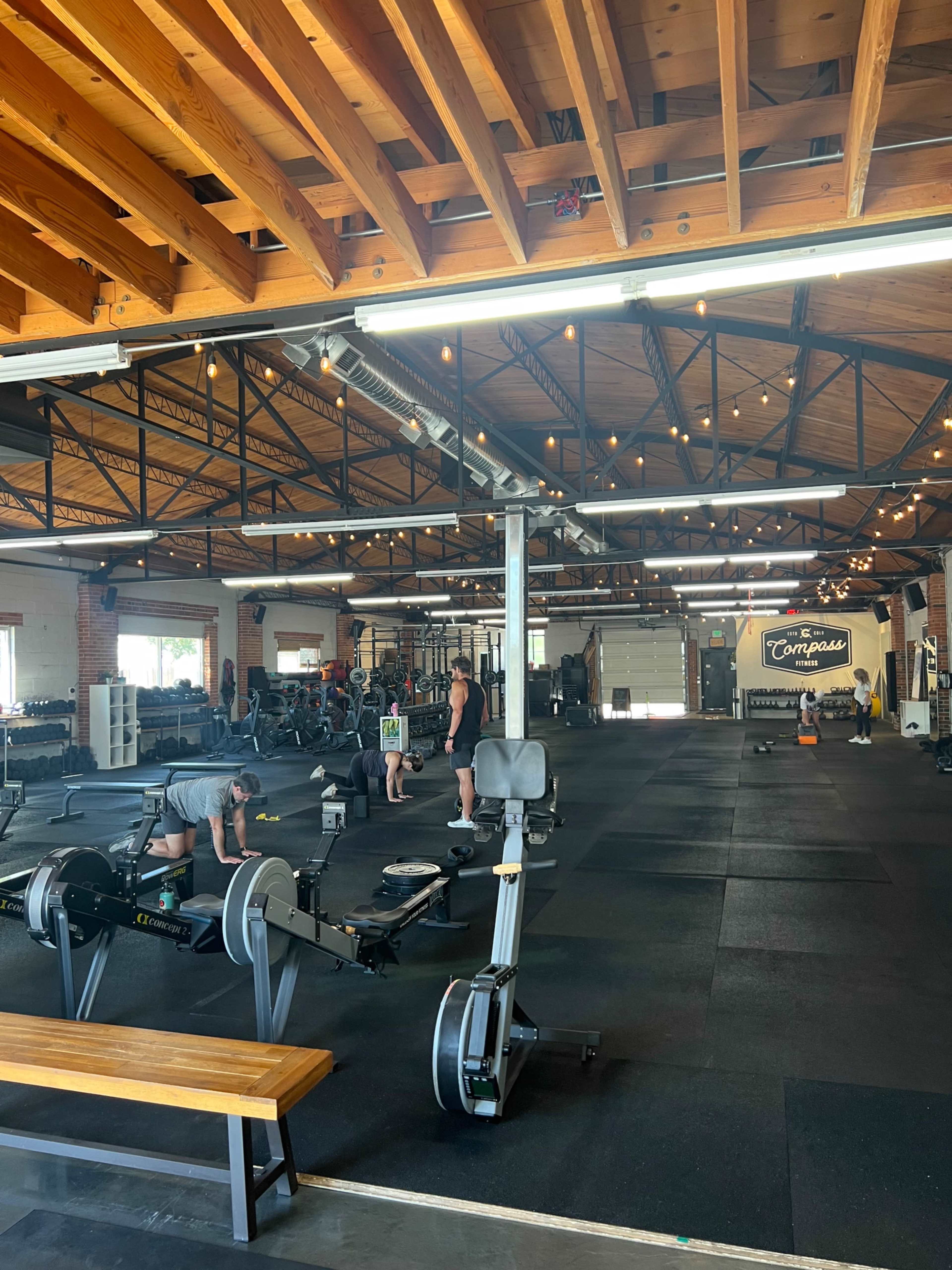 The image shows a spacious gym with various workout equipment, including rowing machines and free weights, under a wooden ceiling with exposed beams.