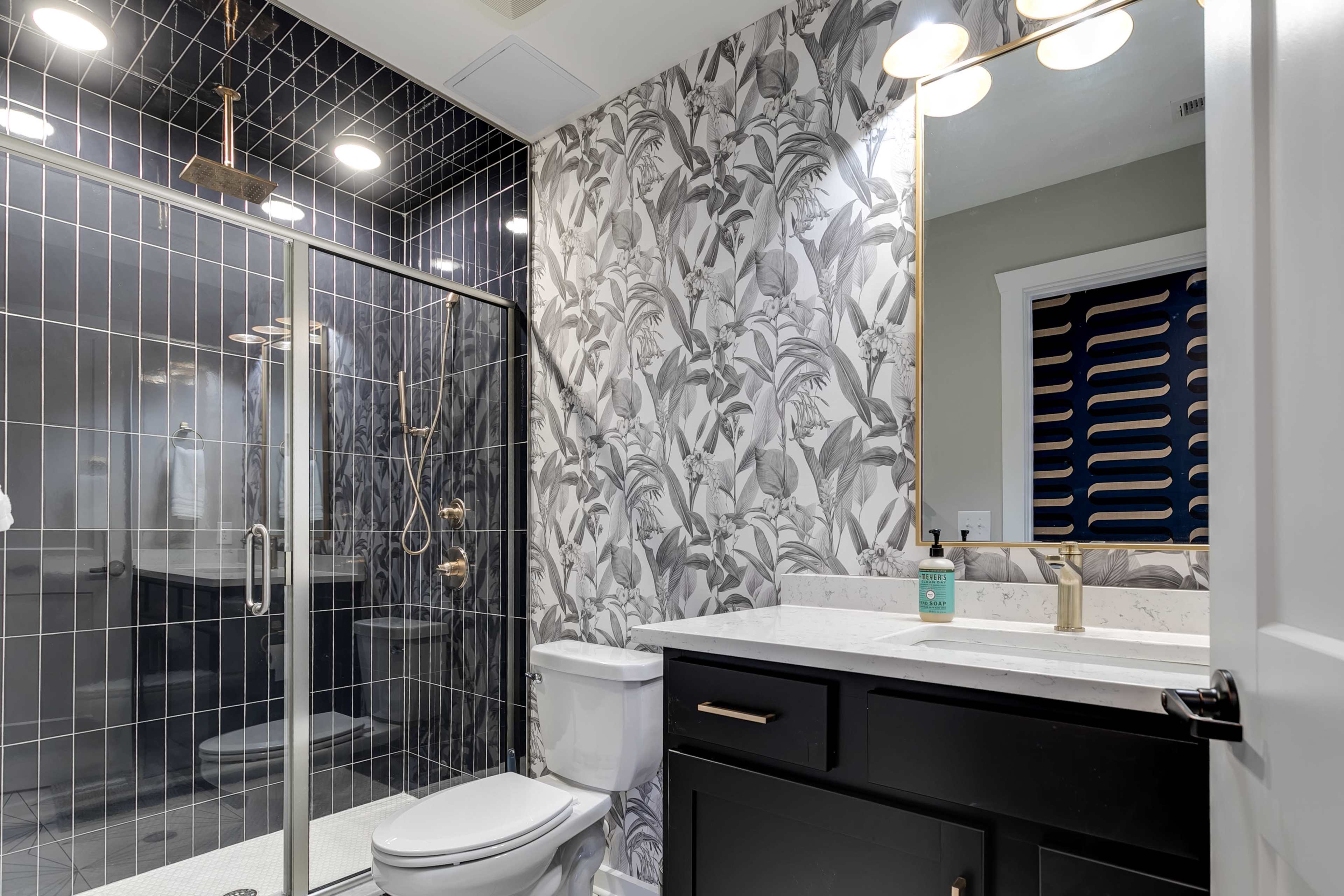 A modern bathroom featuring a glass shower enclosure, a black and white leaf-patterned wallpaper, and a dark cabinet with a white countertop and mirror.