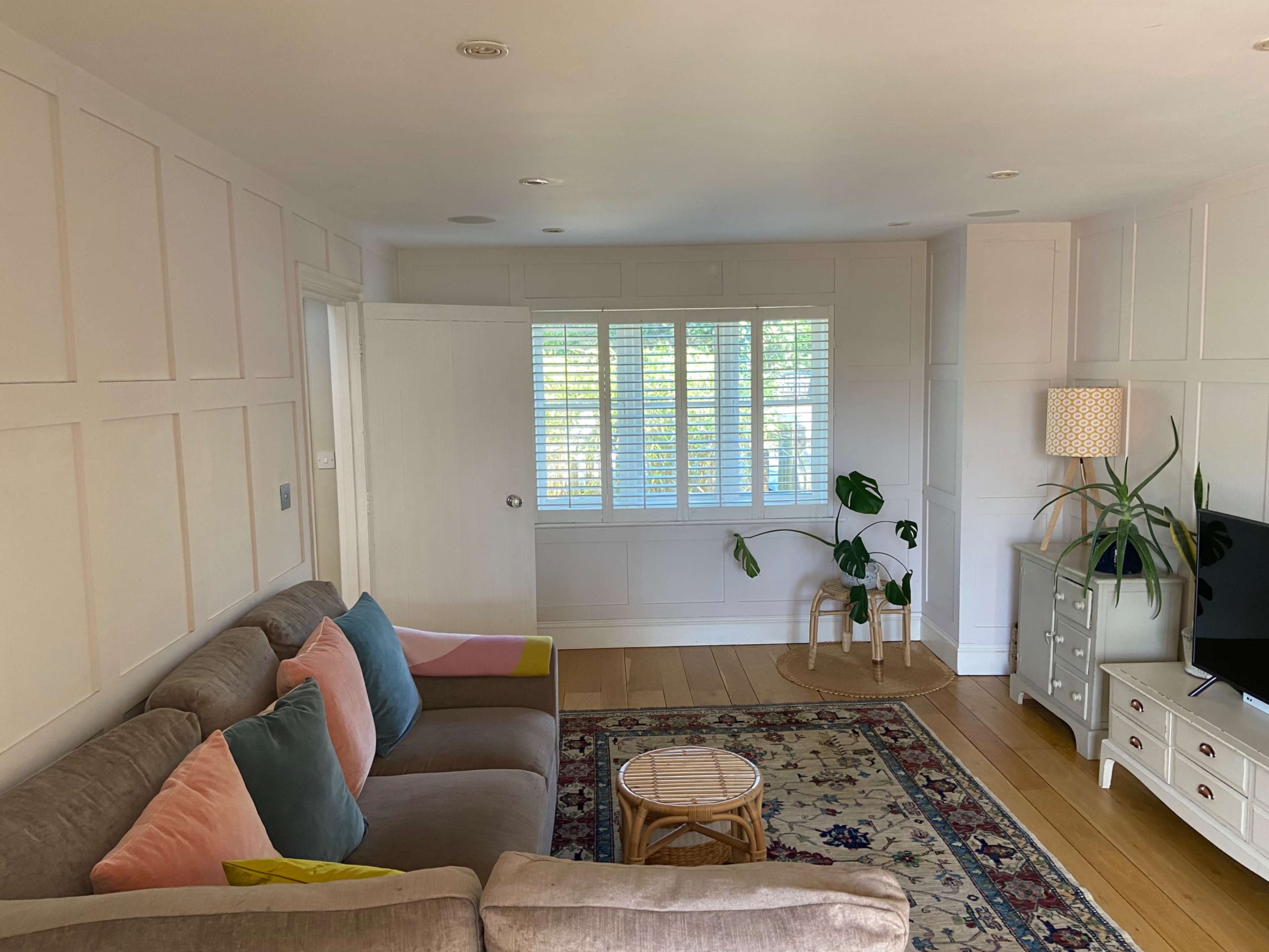 The image shows a living room featuring a beige sofa with colorful cushions, a small round coffee table, and a television on a white cabinet, with natural light coming through shutters.