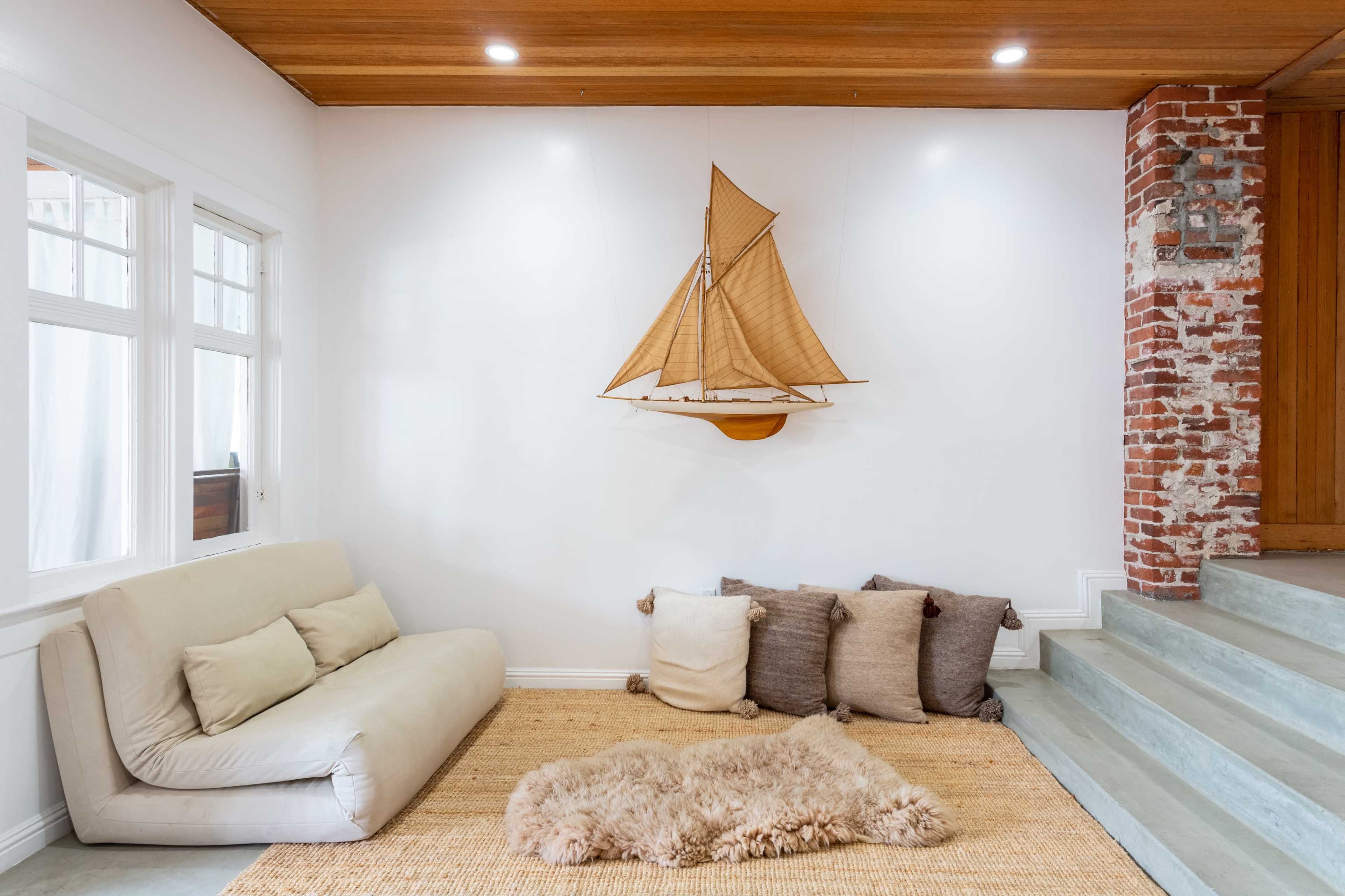 A spacious room features a sailboat model mounted on a white wall, with a light-colored sofa and decorative pillows arranged on a woven rug.