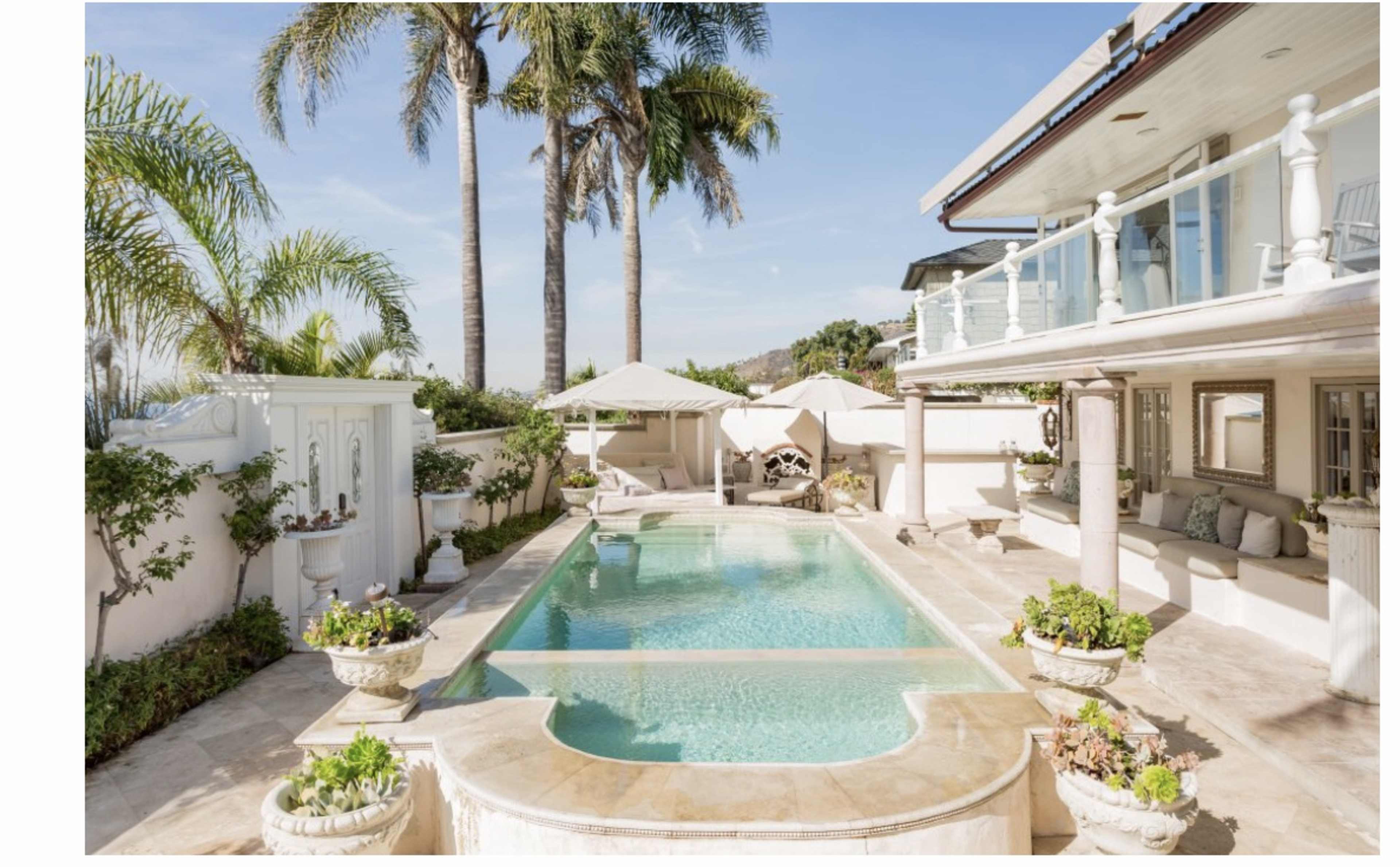A landscaped pool area surrounded by white loungers, palm trees, and a terrace with decorative planters.