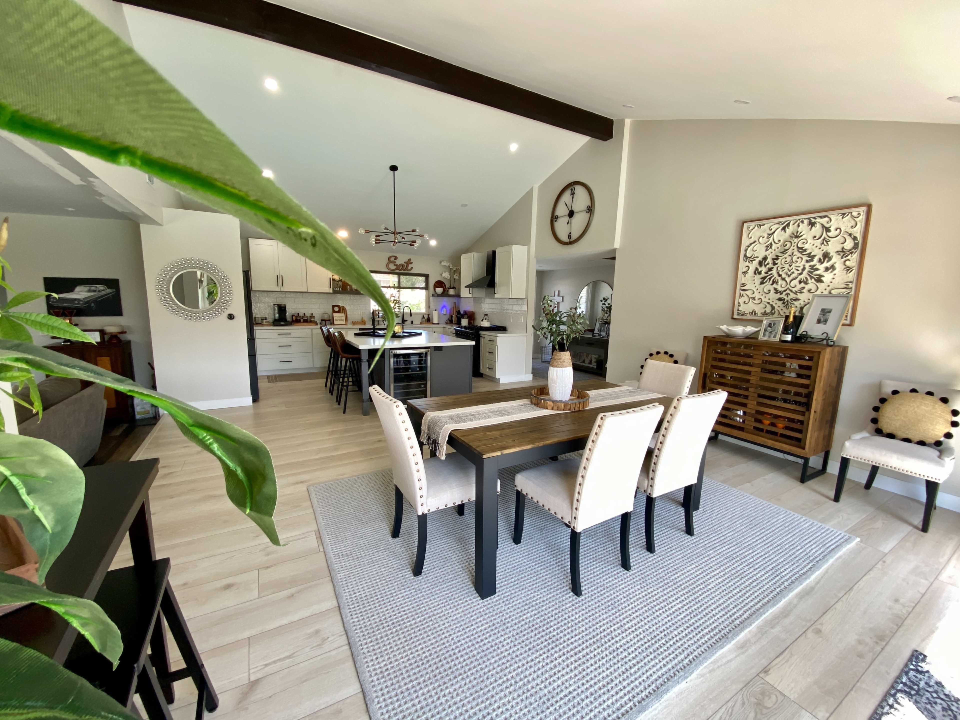 A modern dining area with a long table surrounded by chairs, adjacent to a kitchen featuring white cabinets and a large clock on the wall.
