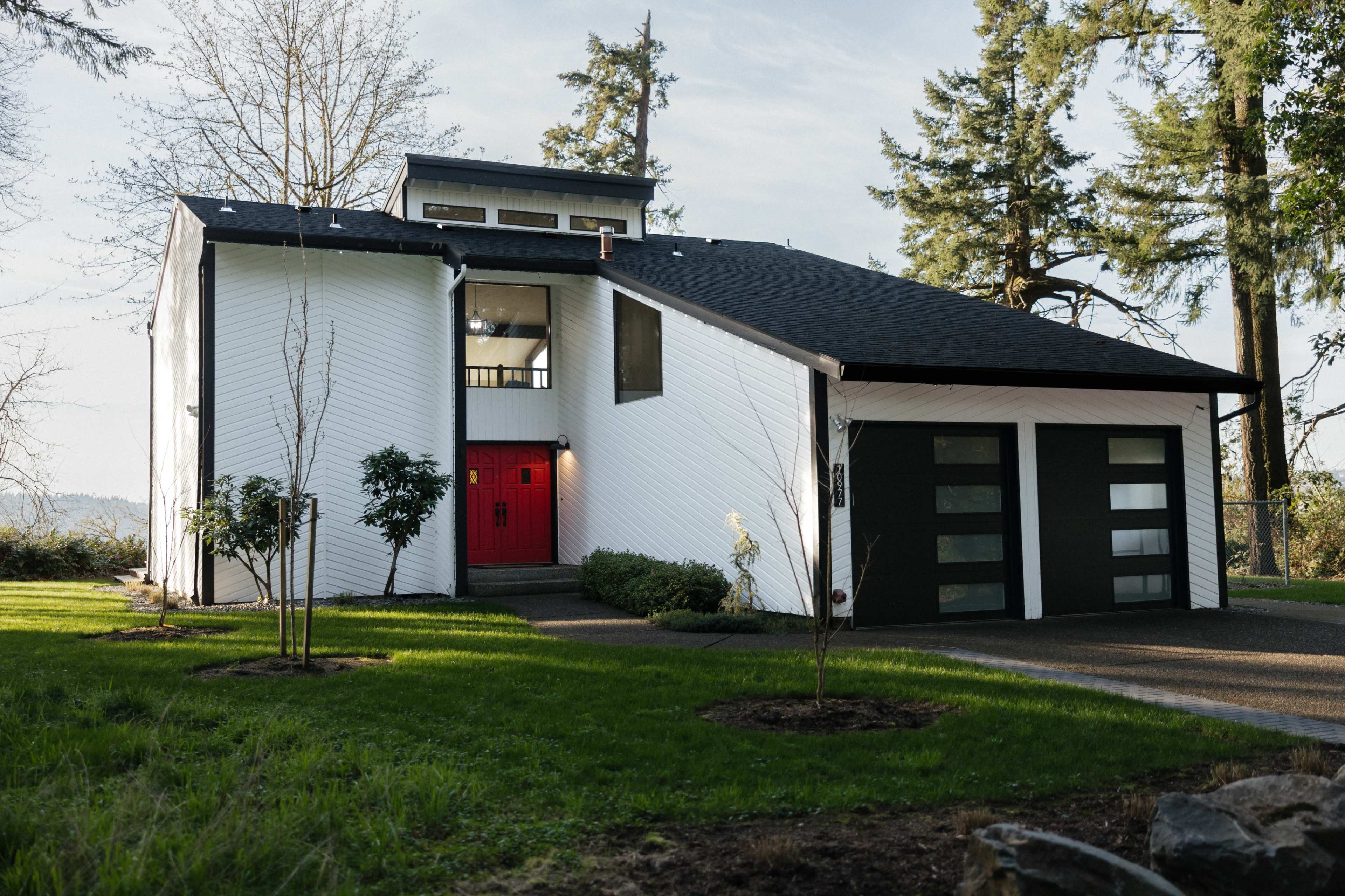 A modern white house with a red front door and large gray garage doors is set amidst greenery and trees.