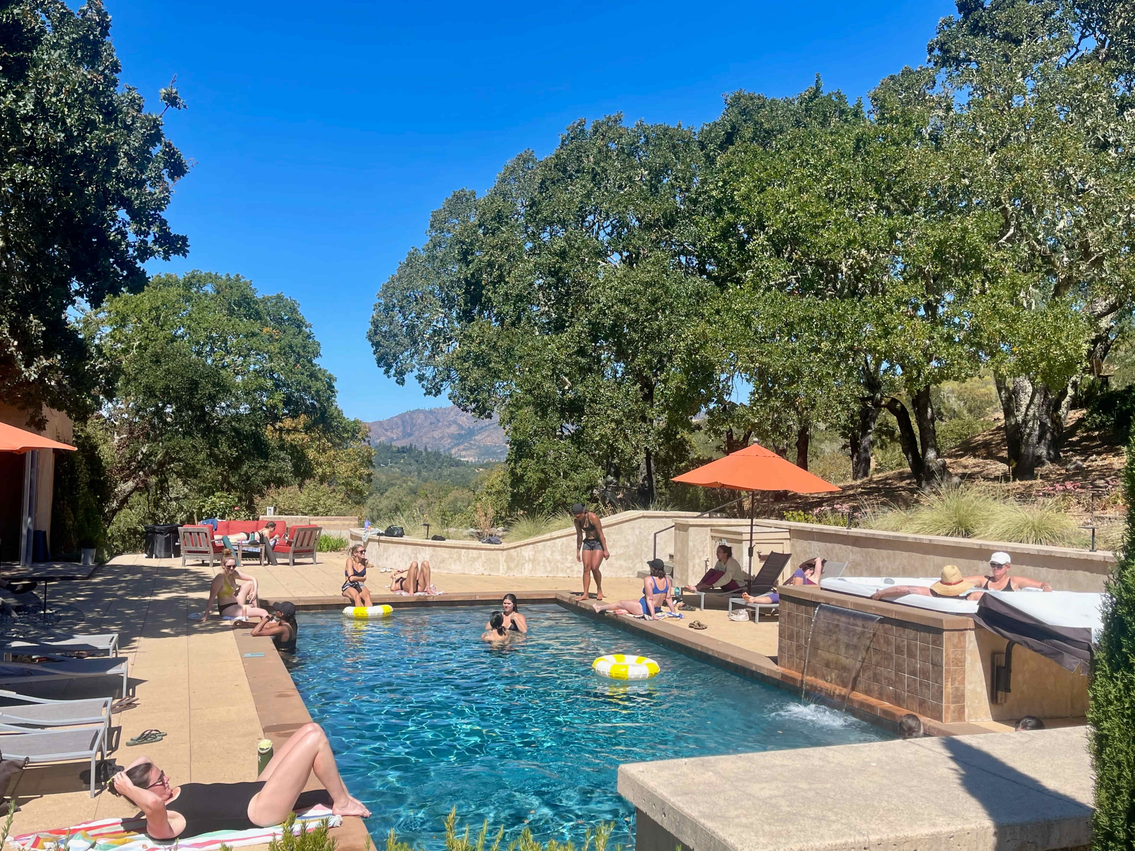 A group of people relaxes by a pool surrounded by trees and mountains under a clear blue sky.