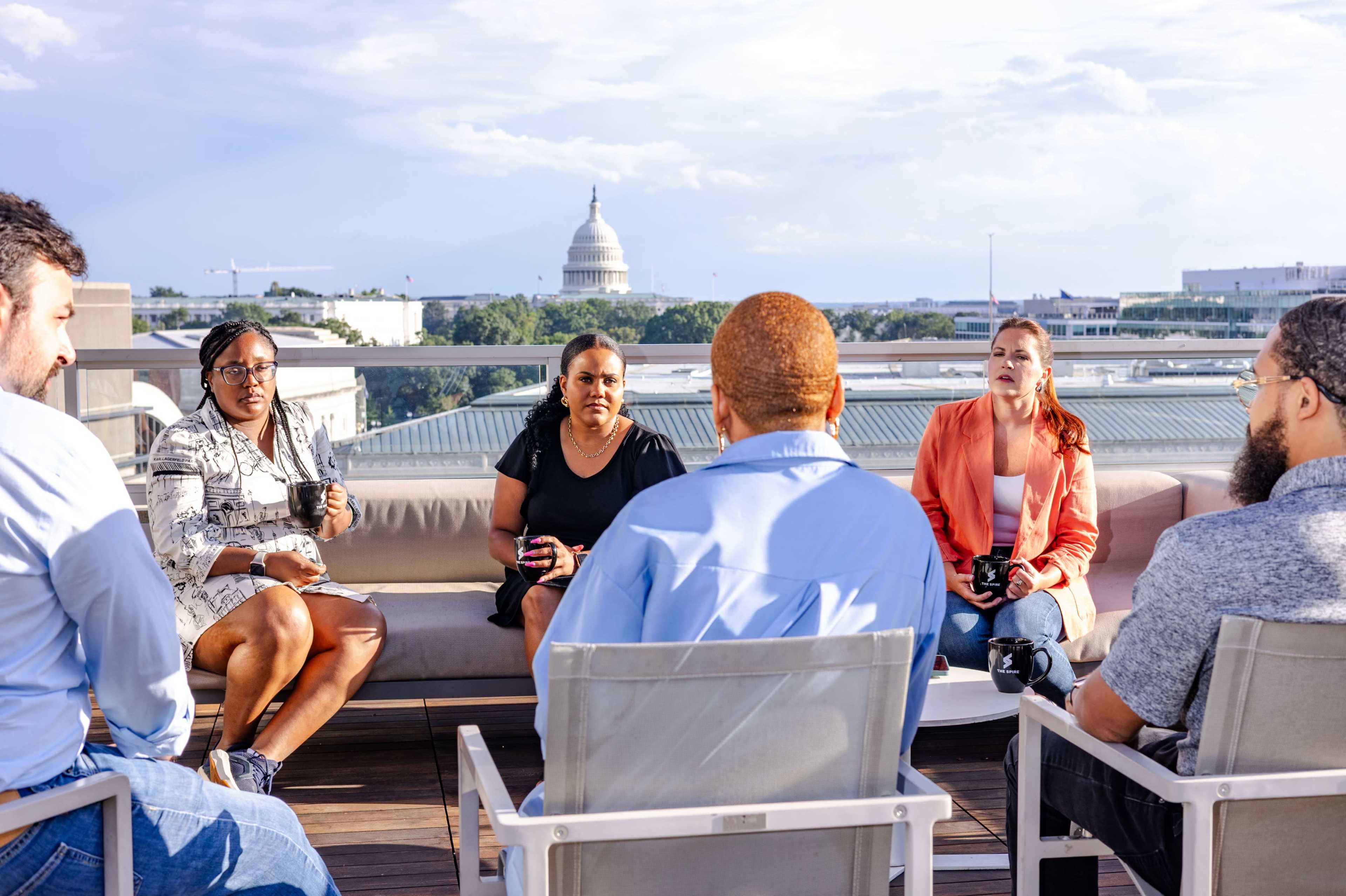 A group of six individuals sits on a rooftop terrace, engaged in conversation, with the U.S. Capitol building visible in the background.