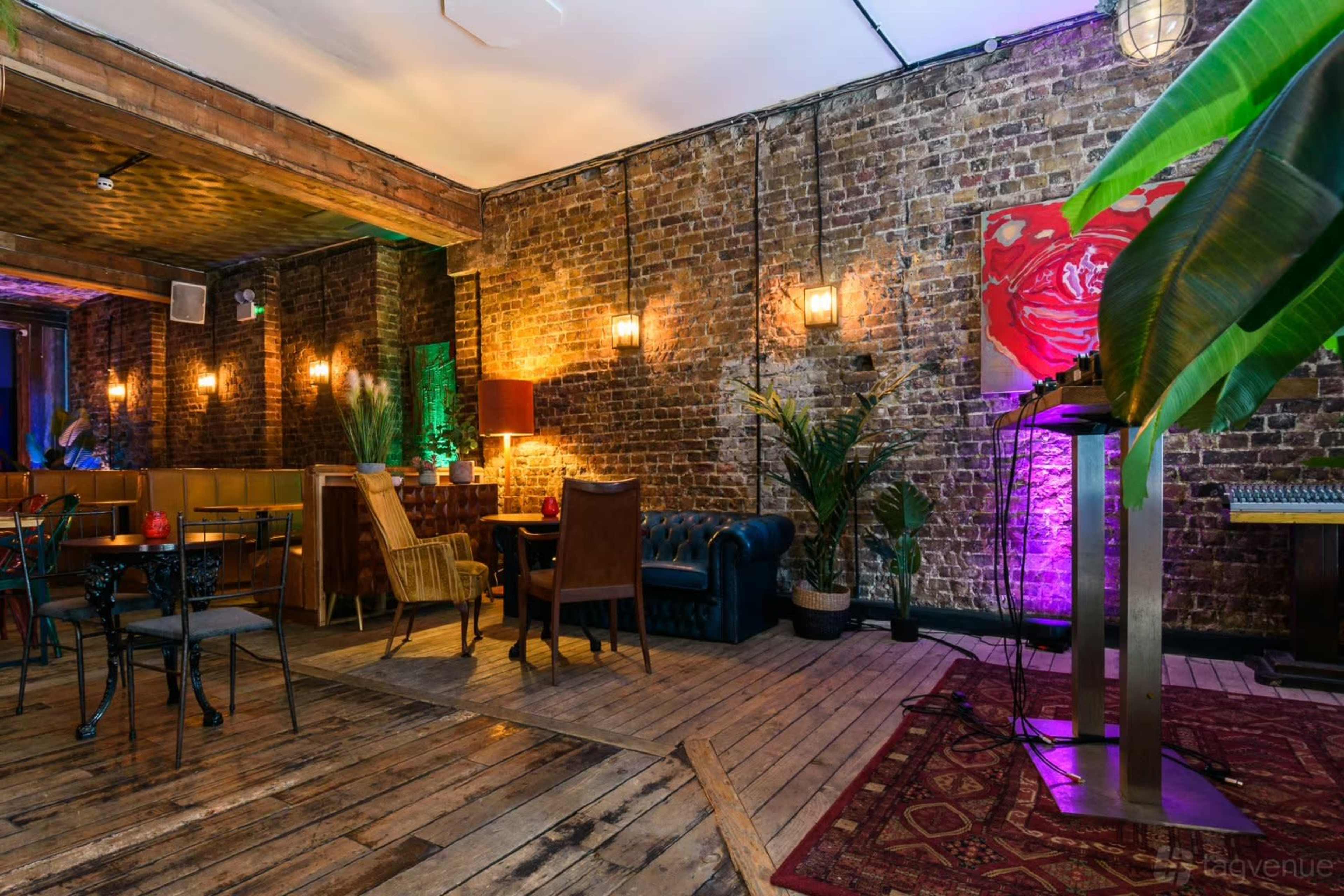 The image displays a rustic indoor space featuring exposed brick walls, wooden floors, and a variety of seating arrangements, including a blue leather sofa and tables with chairs, accented by plants and soft lighting.