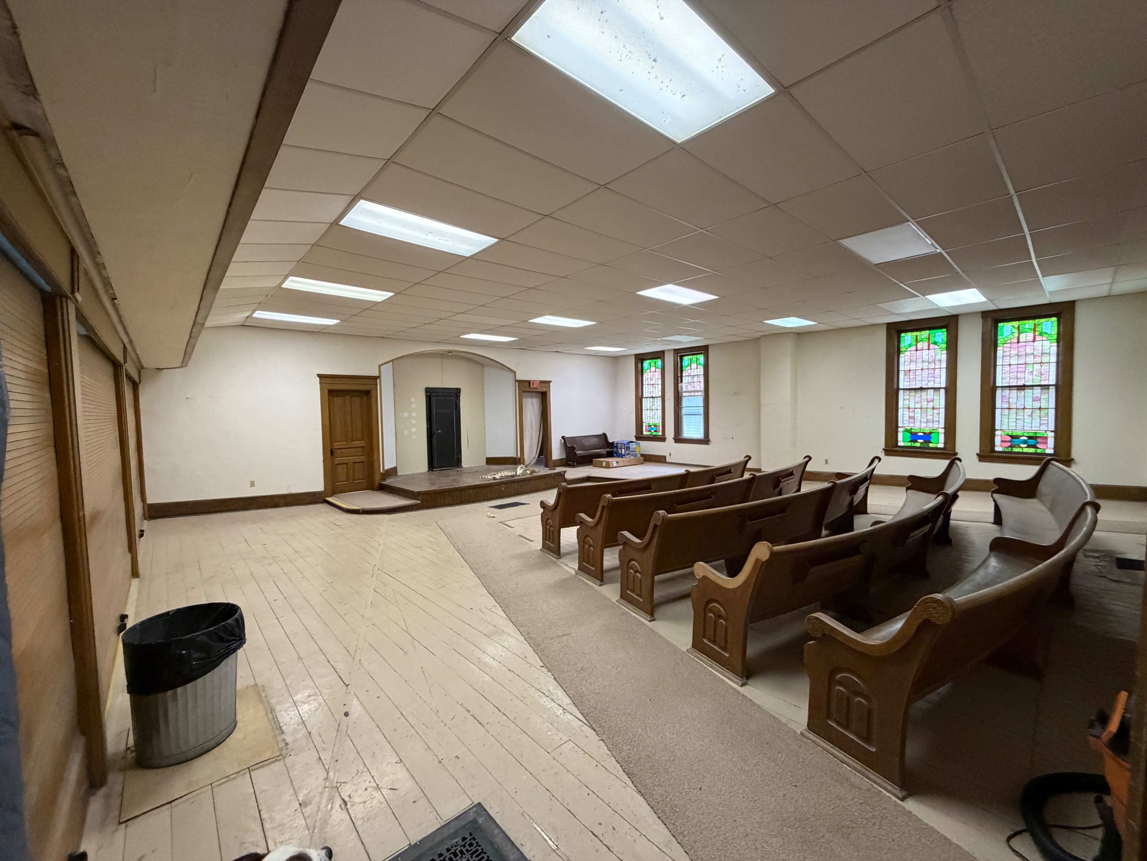 The image shows an empty room with wooden pews, stained glass windows, and a small platform at one end.