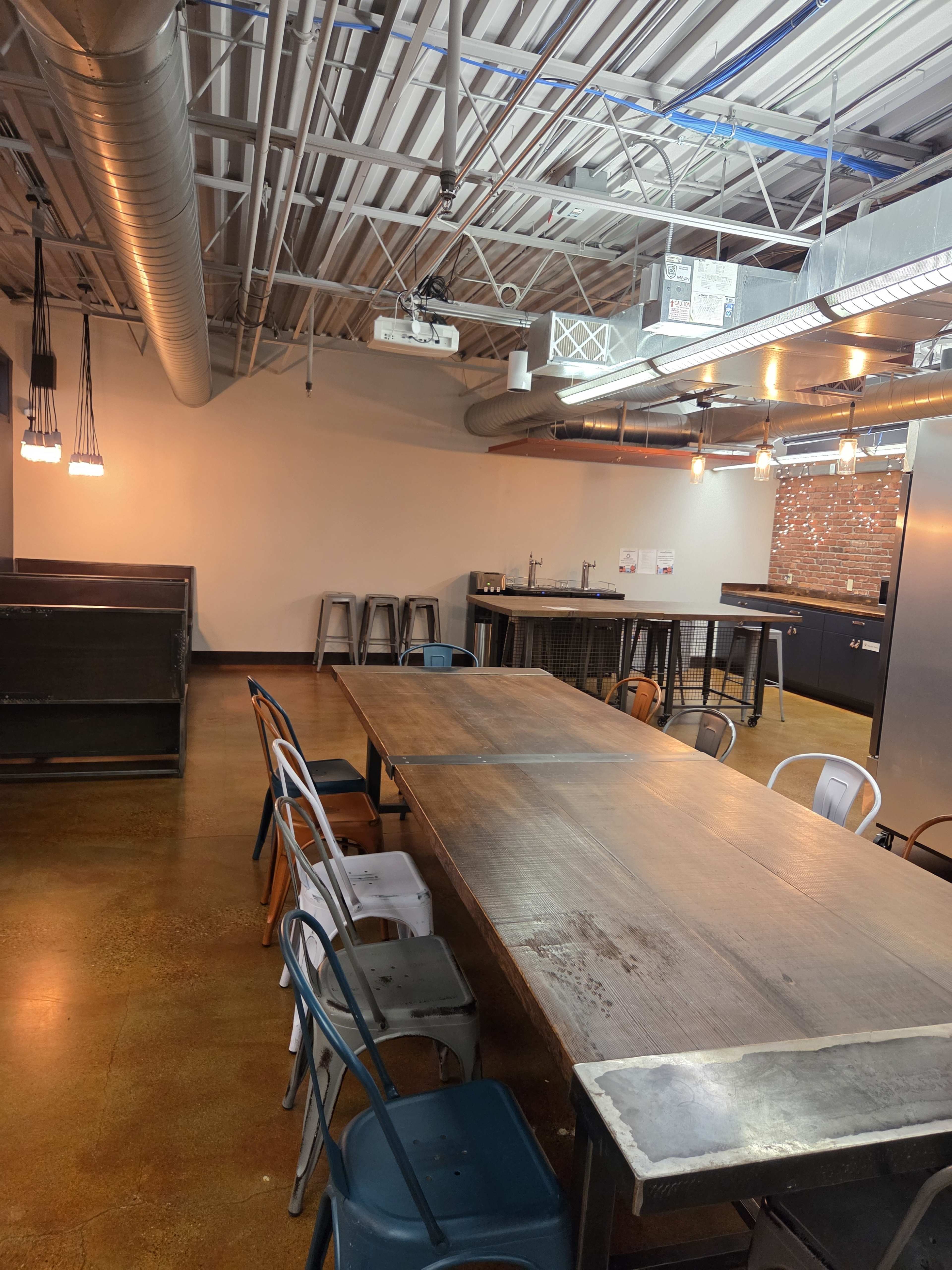 The image shows an industrial-style room with a large wooden table surrounded by various metal and wooden chairs, along with a few high stools, and exposed ductwork and lighting fixtures overhead.