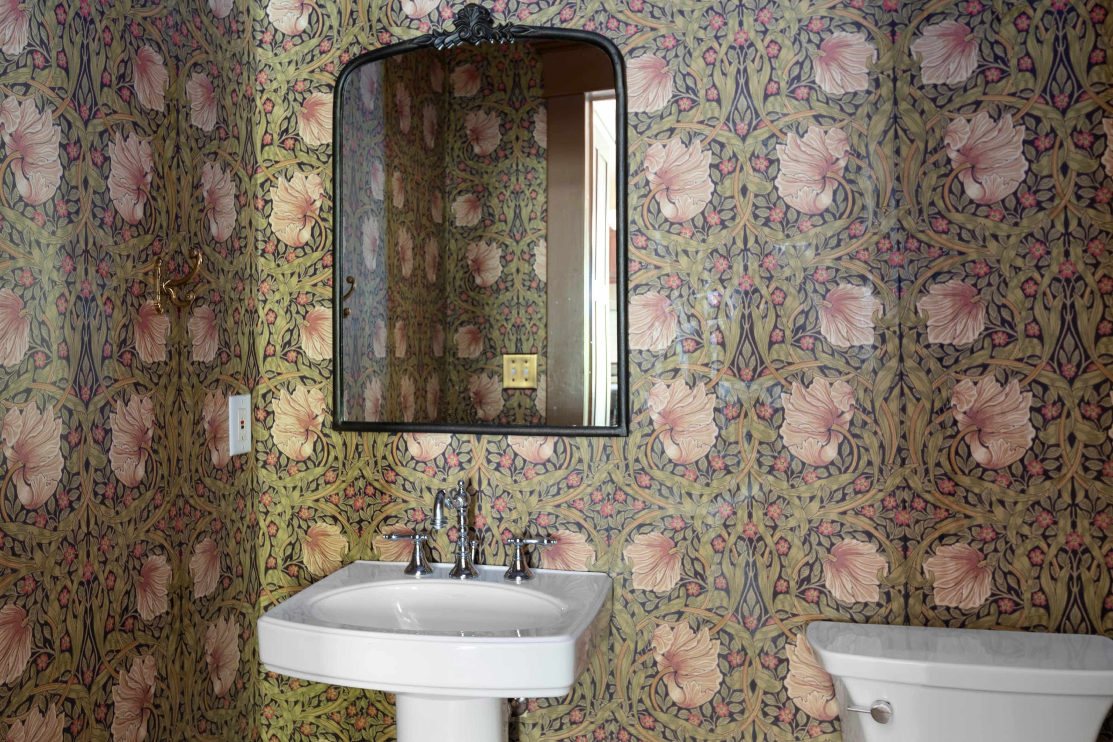 The image shows a bathroom featuring a floral-patterned wallpaper, a white sink with chrome faucets, and a toilet next to the sink.