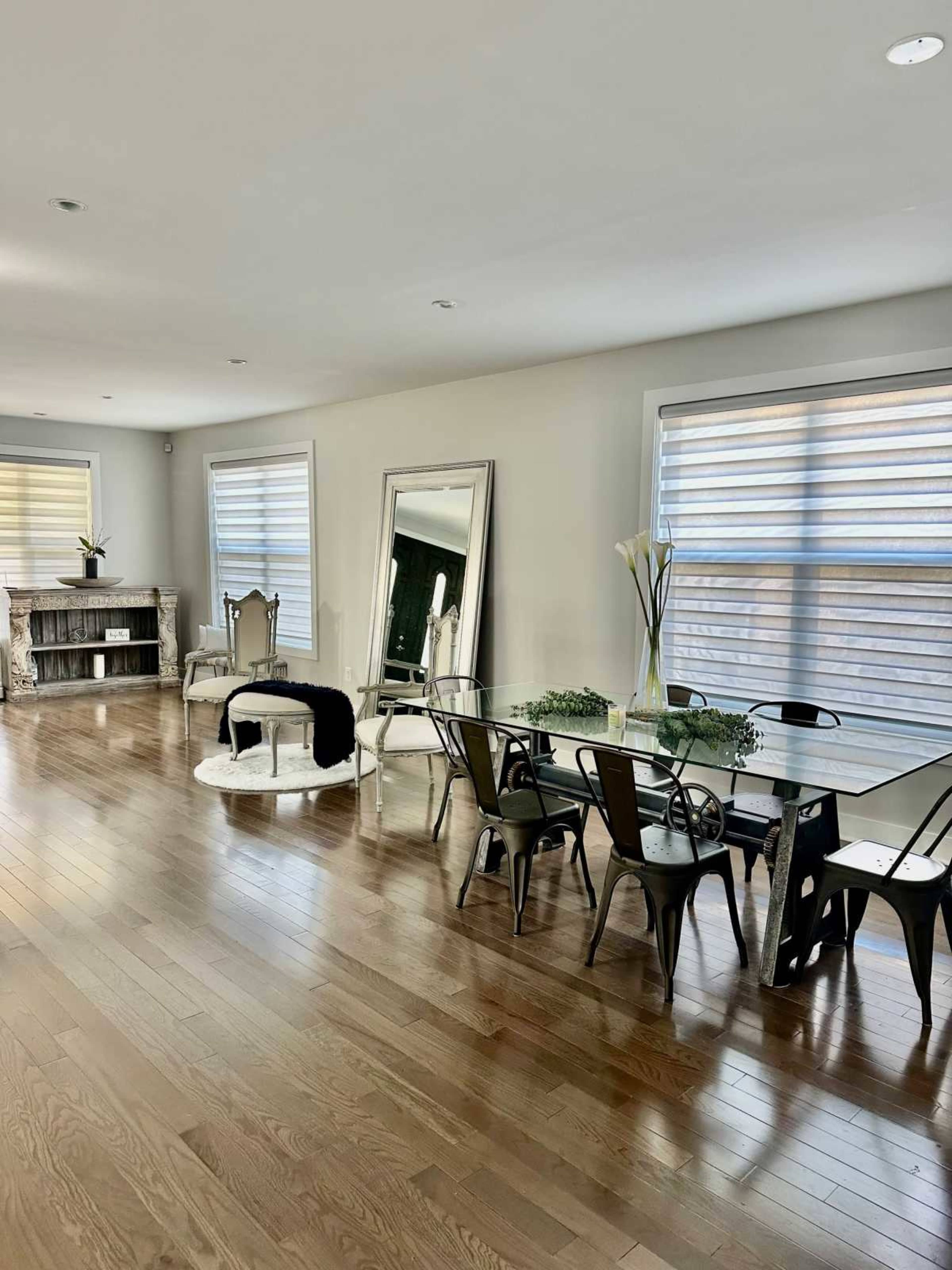 A spacious dining area features a glass table surrounded by metal chairs, with hardwood floors and large windows offering natural light.