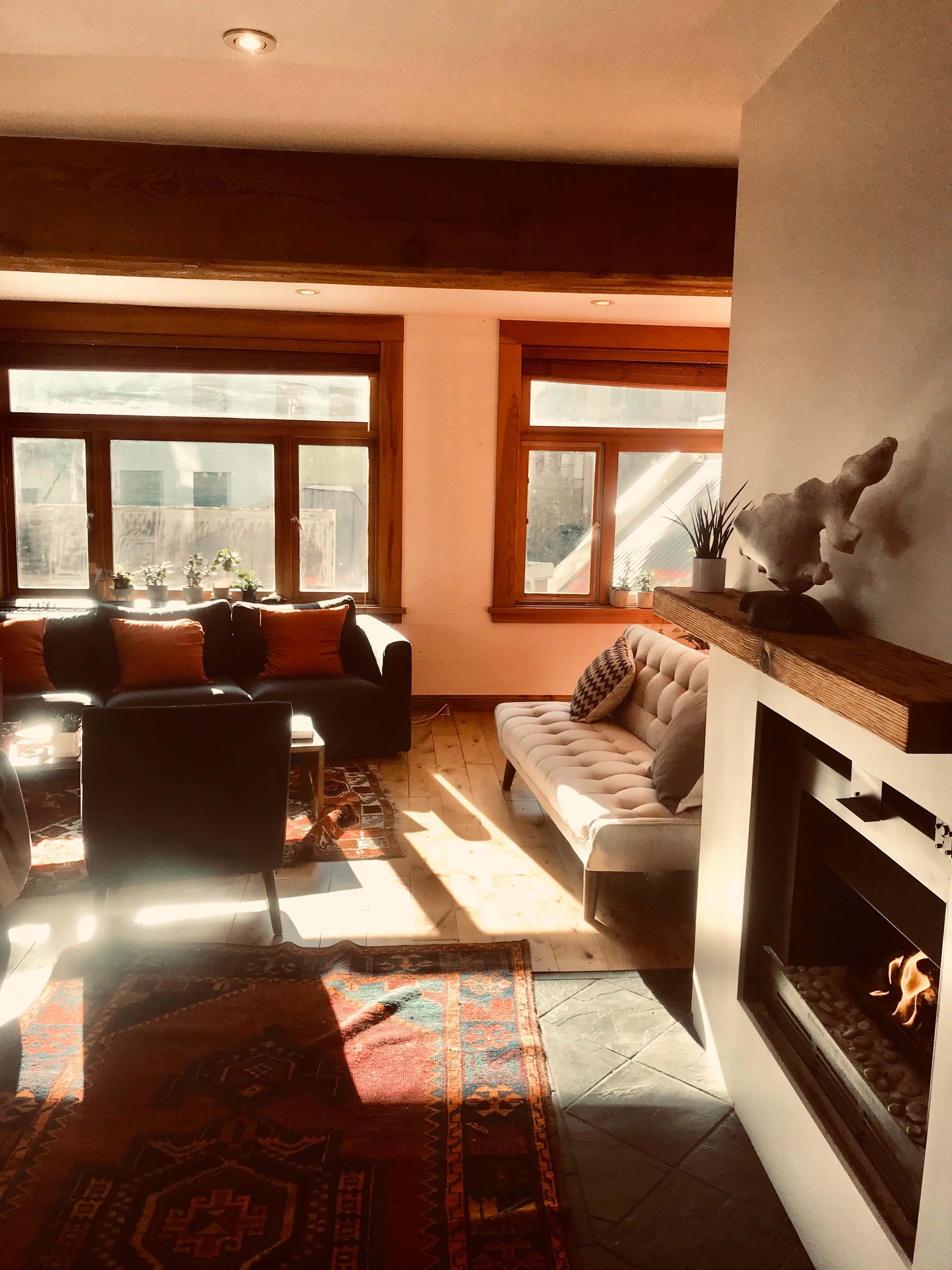 A cozy living room with a light-colored sofa, two black chairs, a fireplace, and large windows letting in natural light.