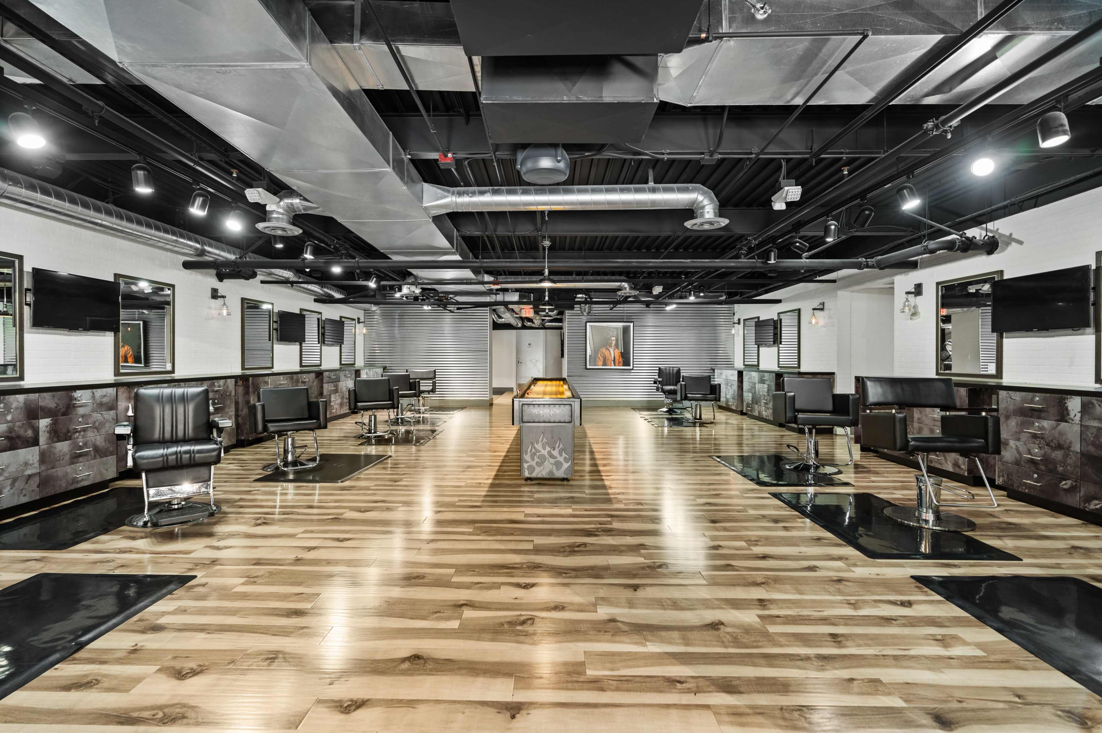 The image shows a modern barbershop interior with multiple styling stations, black chairs, and polished wooden flooring under bright overhead lighting.