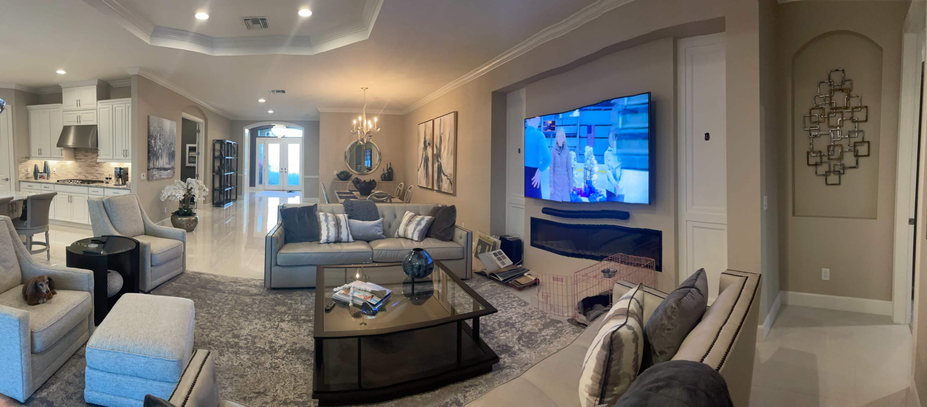 A spacious living room with modern furniture, a large television mounted on the wall, and an open view into a kitchen area.