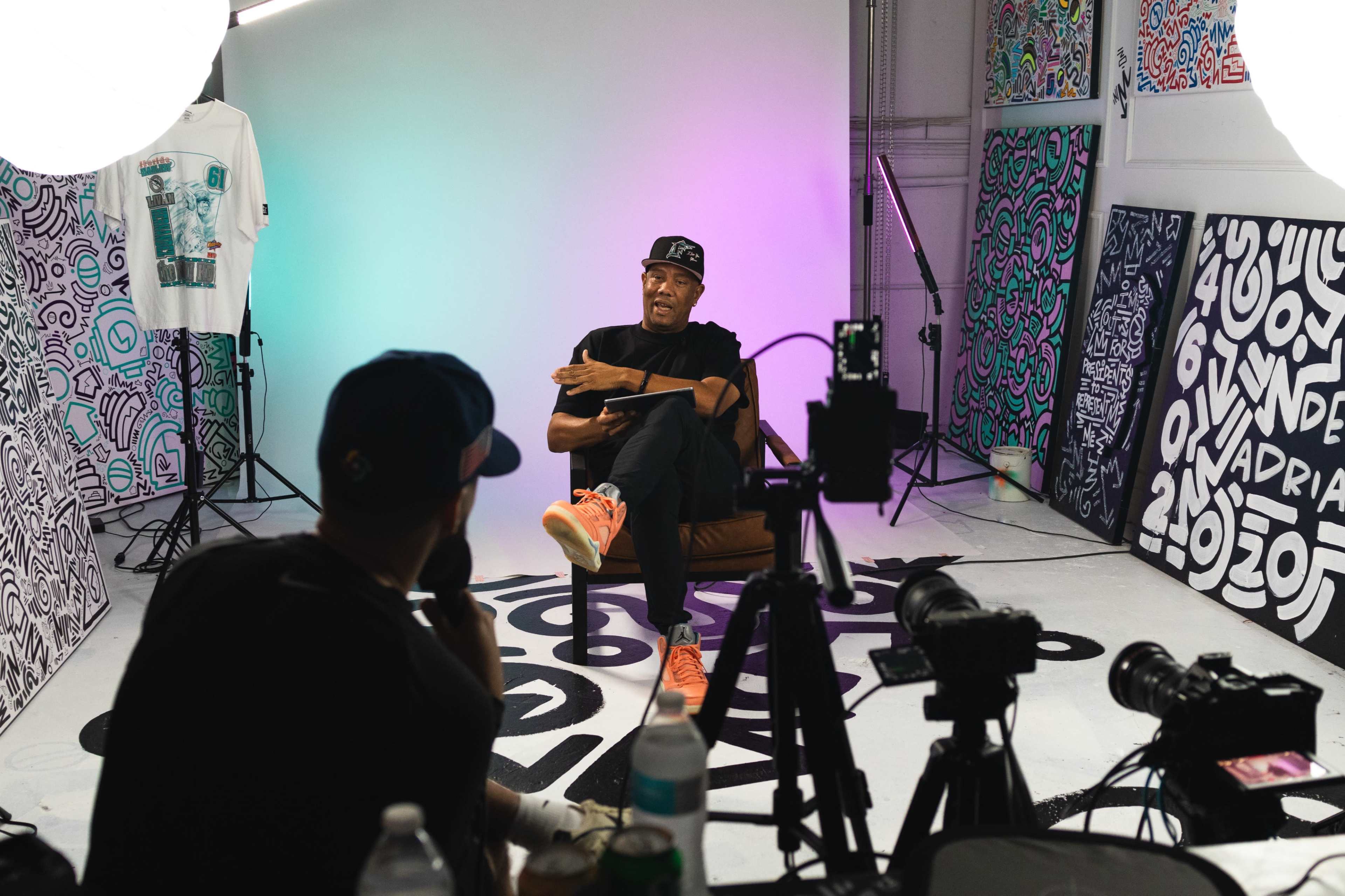 A man sits in a chair, engaged in conversation with another person off-camera, in a brightly lit studio surrounded by colorful artwork and photography equipment.