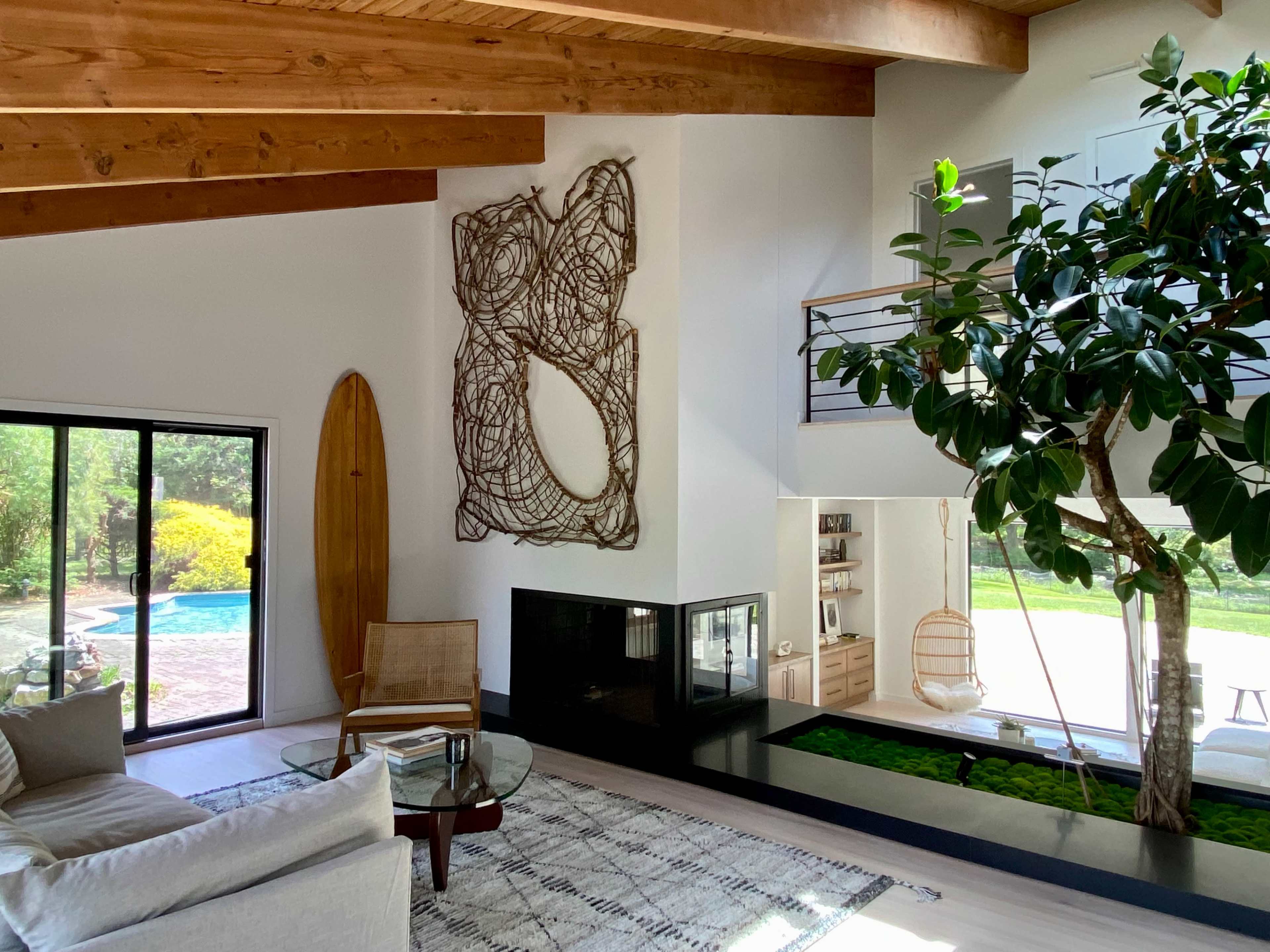 The image shows a modern living room with a large window overlooking a pool, a decorative wall hanging, and a green indoor plant next to a fireplace.