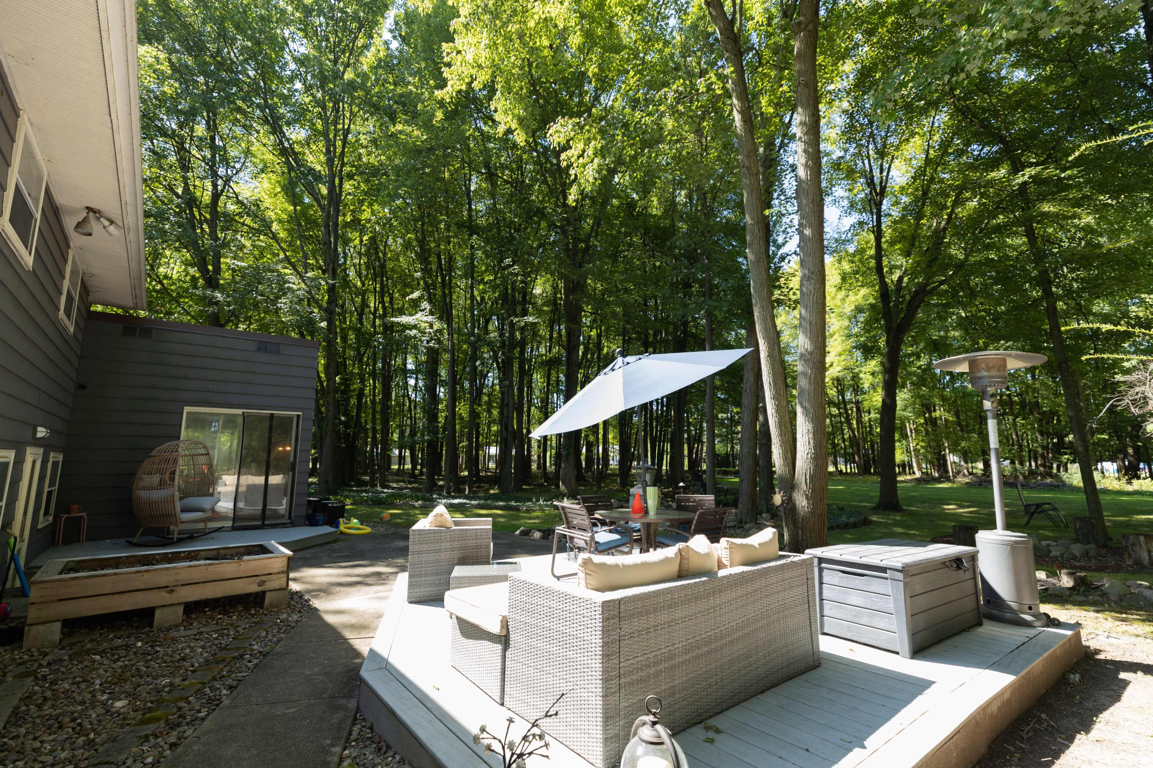 A patio area with outdoor furniture and an umbrella is set among tall trees in a wooded backyard.